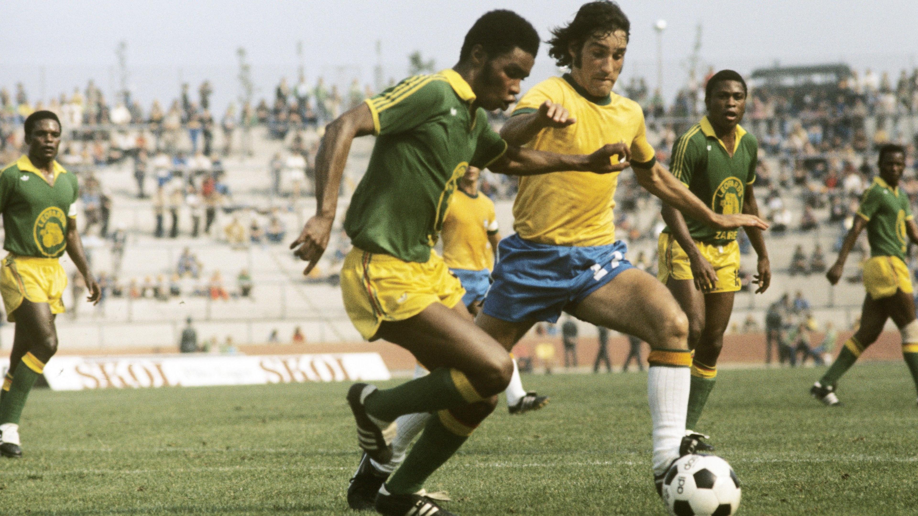 An image from the 1974 World Cup in West Germany, with Zaire taking on Brazil. The central part of the image shows a Zaire player, wearing green socks, yellow shorts and a green shirt with yellow trim, tussling for a black and white ball against a Brazilian opponent, who is wearing white socks, blue shorts and a yellow shirt. The Zaire player is pushing out his left arm at chest level in a bid to ward off the tackle. Three more Zaire players can clearly be seen watching on in the background, while behind them can be seen a relatively sparsely populated concrete stand