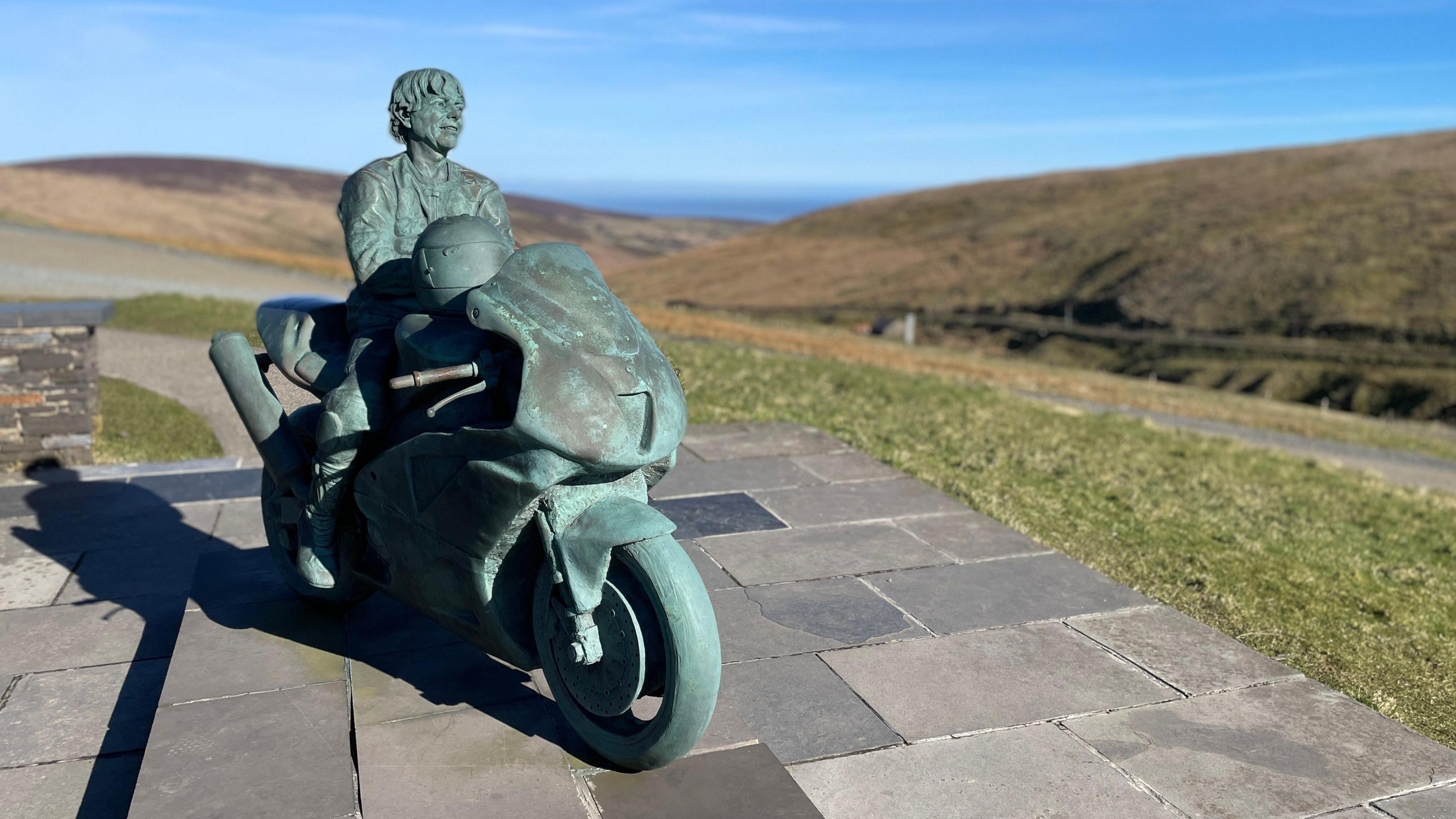 A green statue of Joey Dunlop sitting on a motorcycle while holding a helmet. The statue sits on a paved area next to some green grass by the Mountain Course.