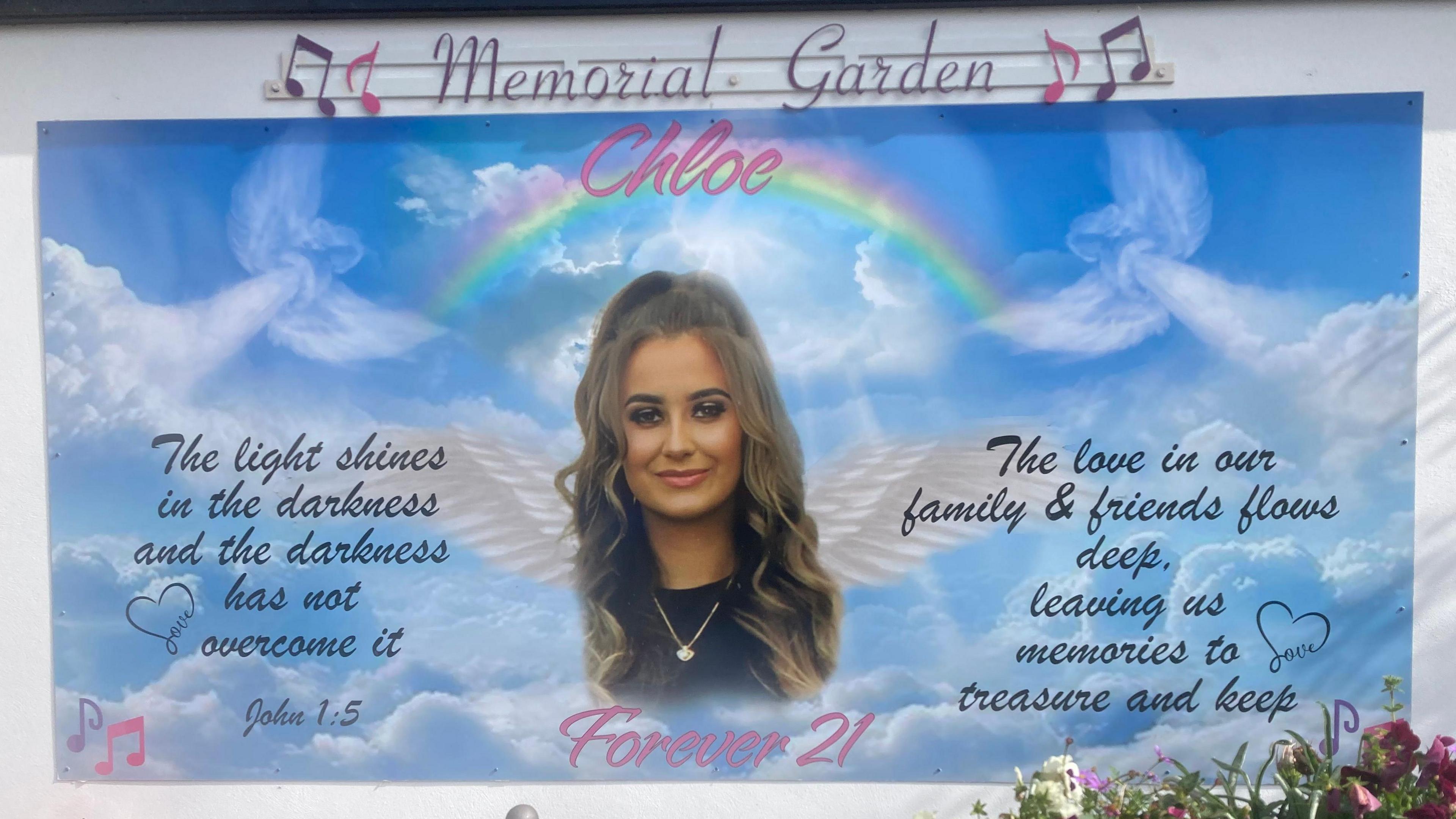 A memorial garden for Chloe.

Her face is in the centre, she has long brown curly hair and is wearing foundation, mascara and lipstick. She is wearing a fine necklace and is smiling with her mouth closed. 

She has white wings coming from her just below her face.

There is a rainbow above her with her name 'Chloe' above her face and then 'Forever 21' below her face. 

On the left hand side of her face, in black writing reads John 1:5 'The light shines in the darkness and the darkness has not overcome it.'

On the right hand side of her face, in black writing reads 'The love in our family & friends flows deep, leaving us memories to treasure and keep'. 

The background is a blue sky with clouds and angels subtly drawn and black hearts and pink music notes. 