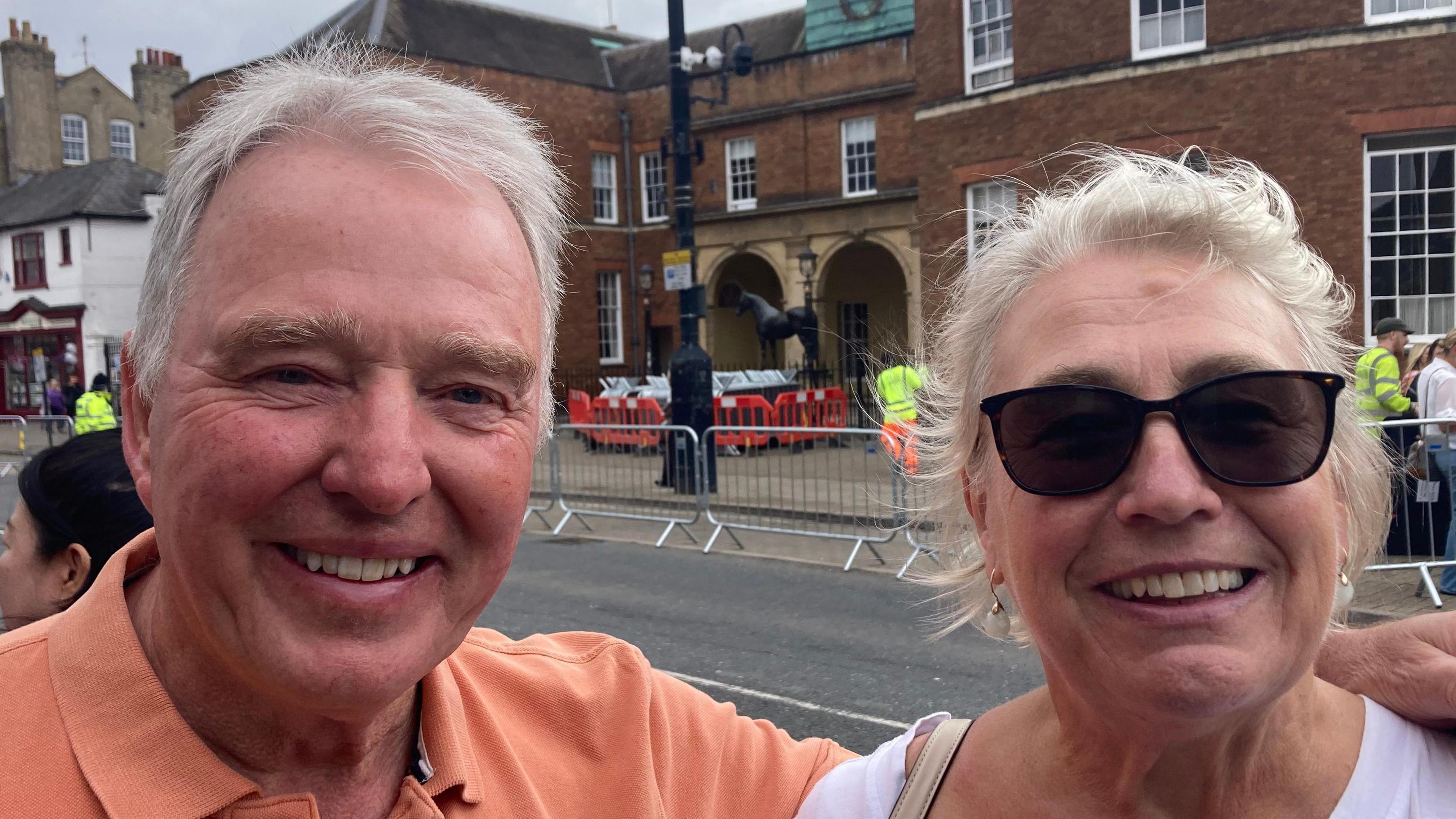 Robert and Robina Hicks smile at the camera. Robert has short grey hair and wears an orange polo top. Robina has grey hair that is tied back and she wears black sunglasses and a white top. 