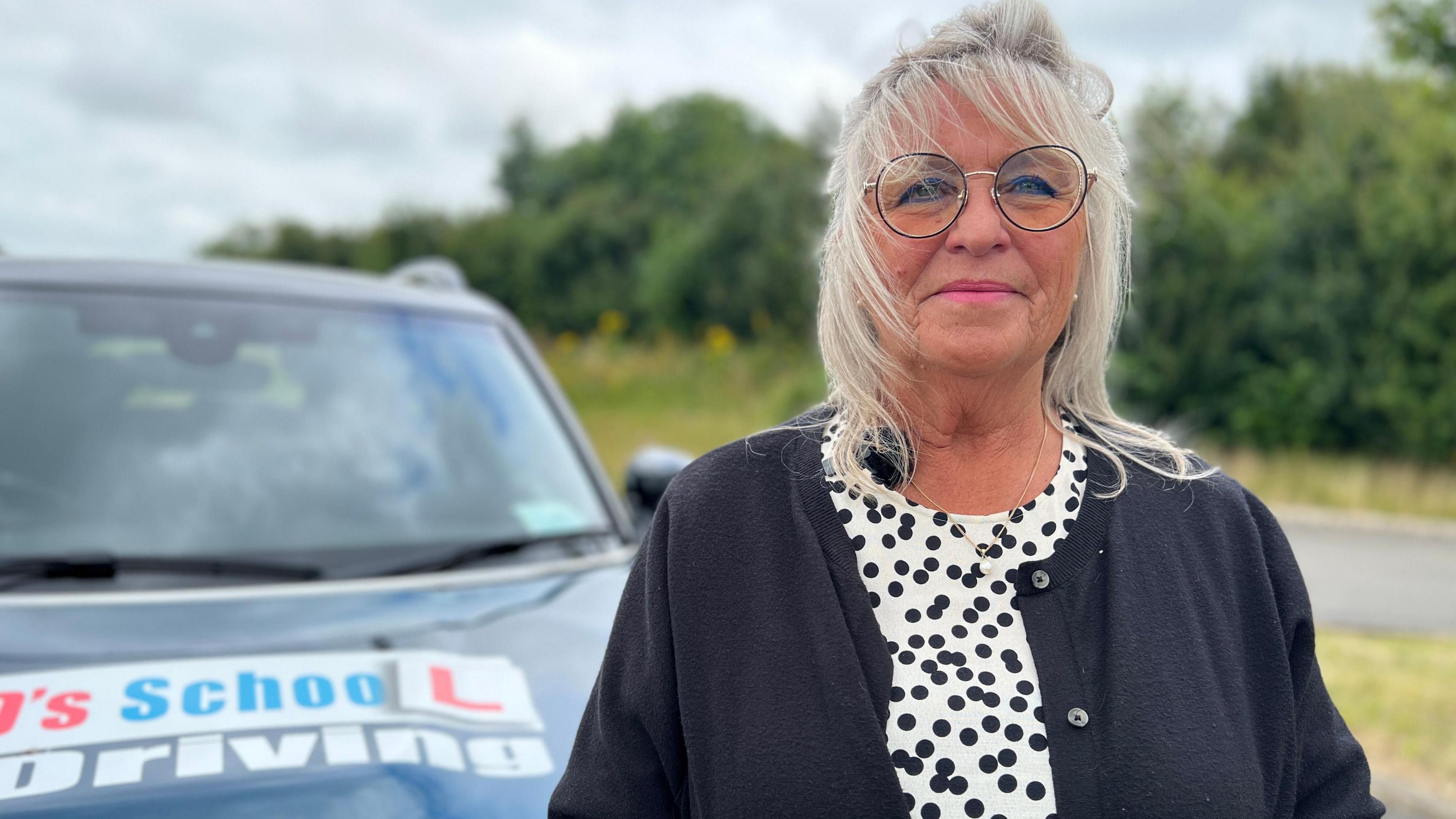 Sue Papworth looking directly at the camera dressed in a white and black spotty dress and black cardigan, with a driving school car behind her