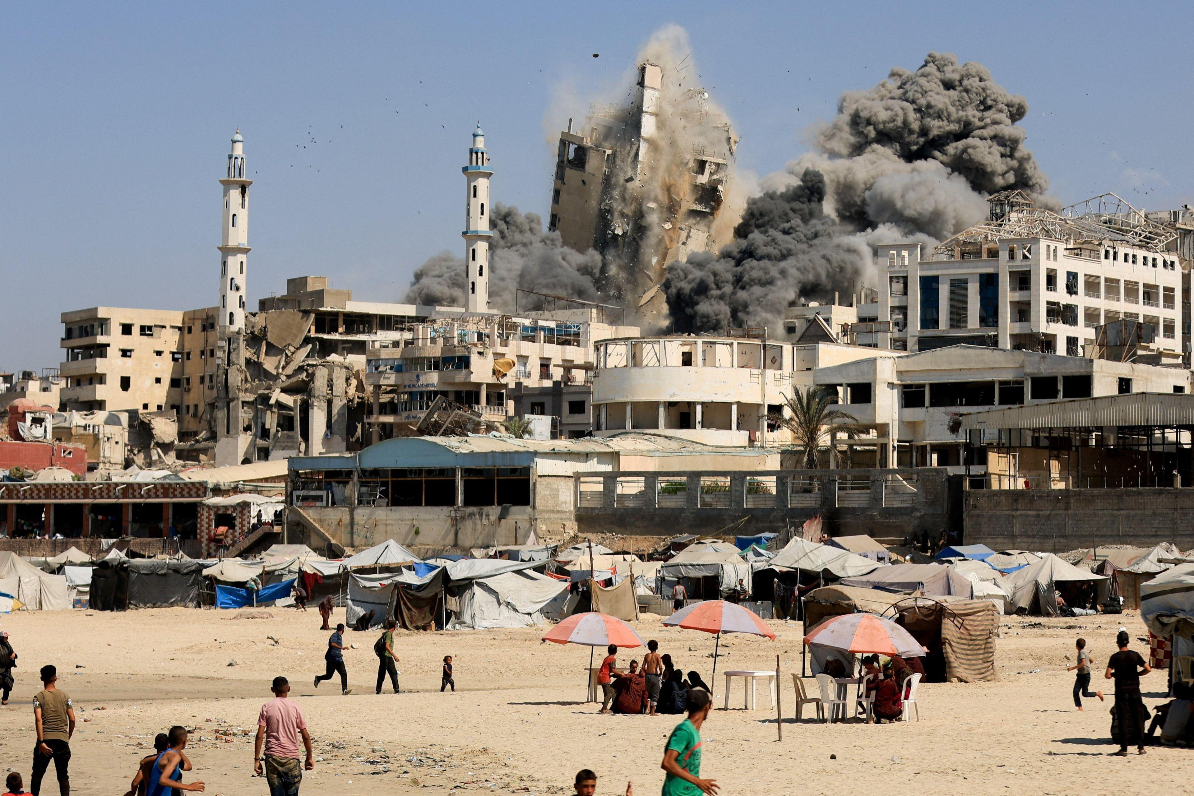 A tower block in Gaza City collapses after being hit by an Israeli air strike. Black smoke rises, and people on a beach run away