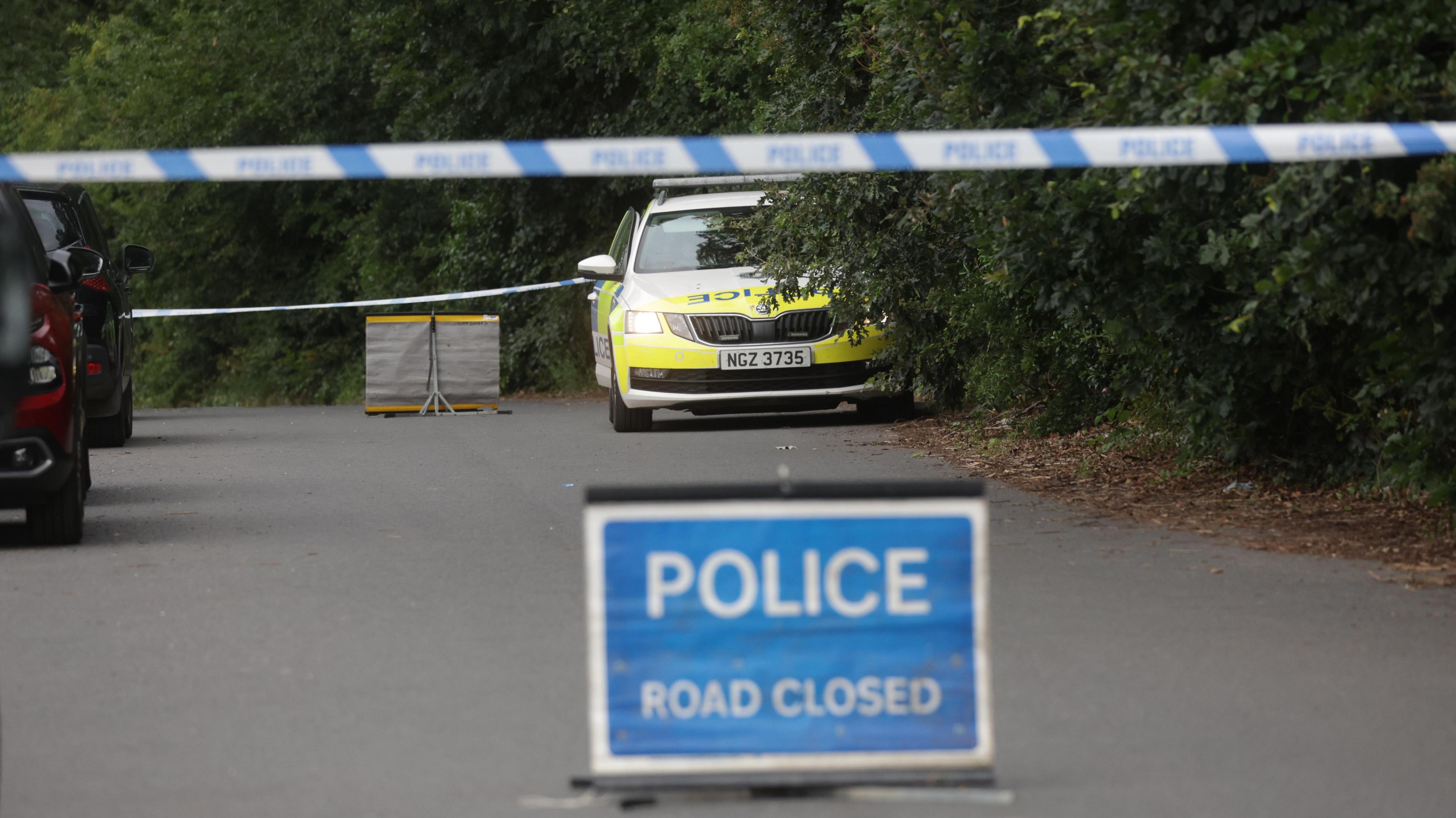 A Skoda police car is parked on a road beside overgrown hedges. There is blue and white police tape across the road and a blue and white police sign saying "POLICE road closed".