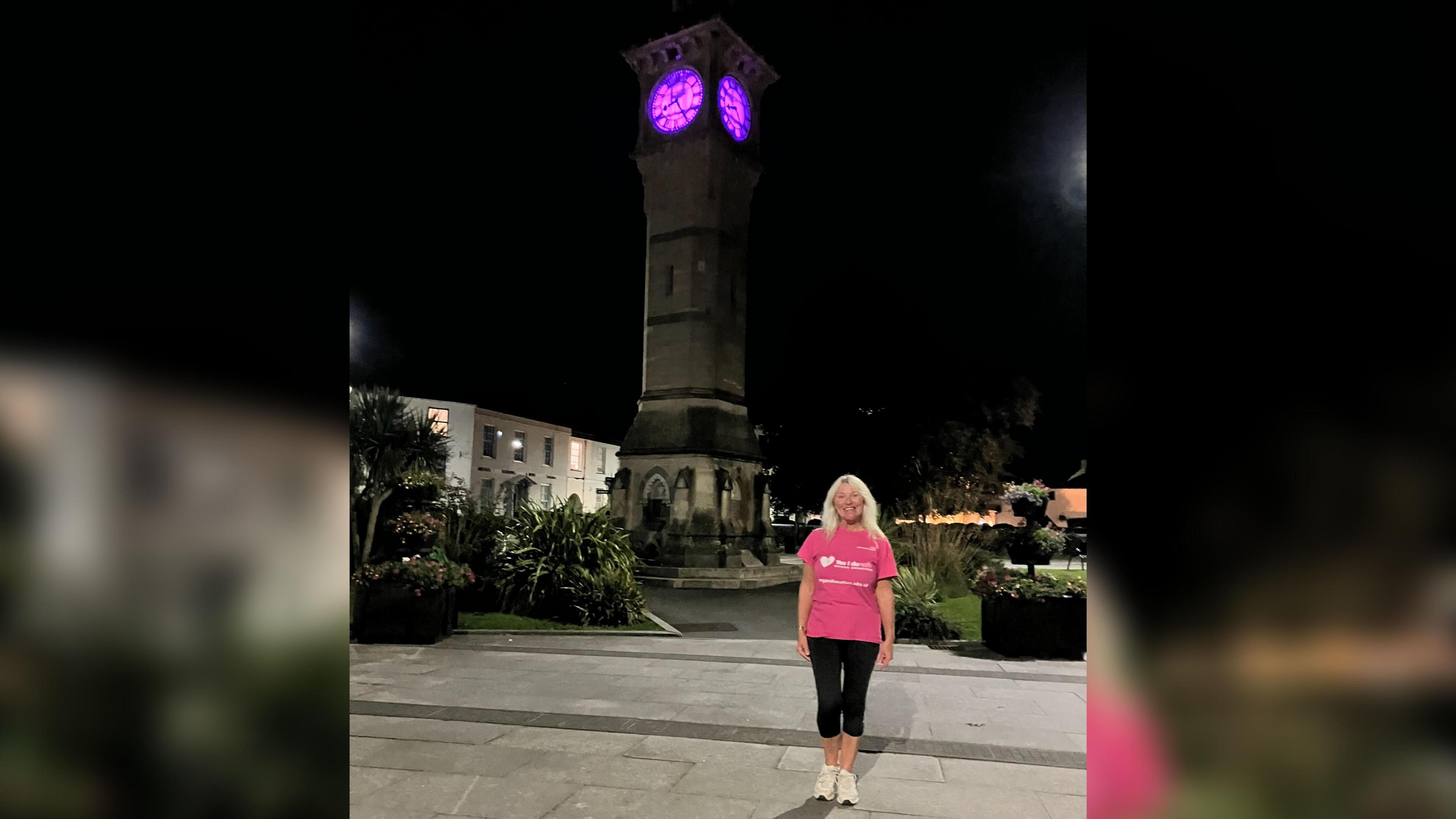 Lisa Wilson is smiling as she stands in front of the clock. It is lit up in pink. She has long blonde hair and is wearing a pink organ donation T-shirt. 