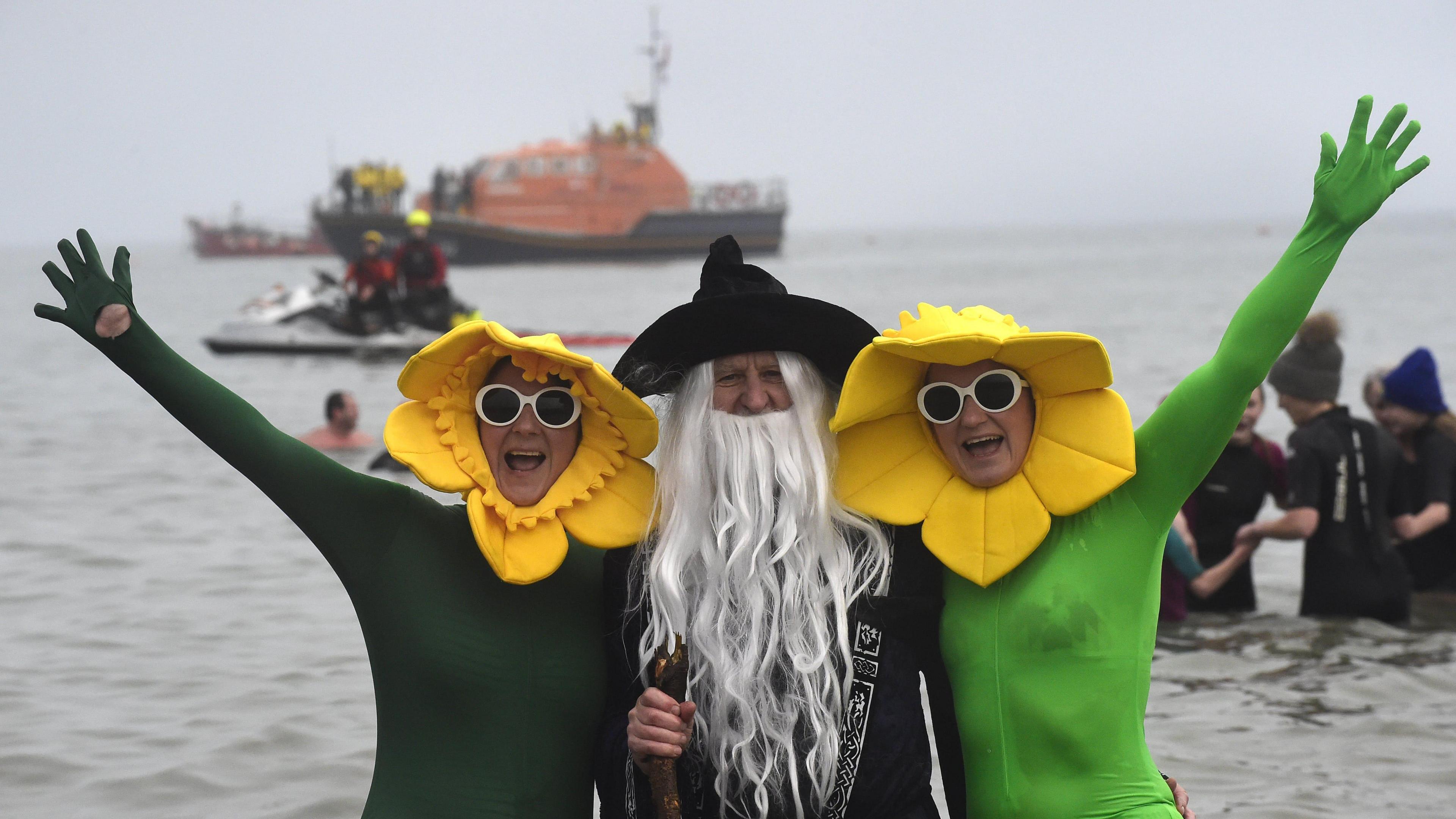 Swimmers in fancy dress during the 52nd Boxing Day swim on December 26, 2024 in Tenby, Wales. Two people in green morph suits with daffodil hats on and wearing white glasses. Between them is a person dressed as a wizard with a long grey beard and black hat and cape and a staff.