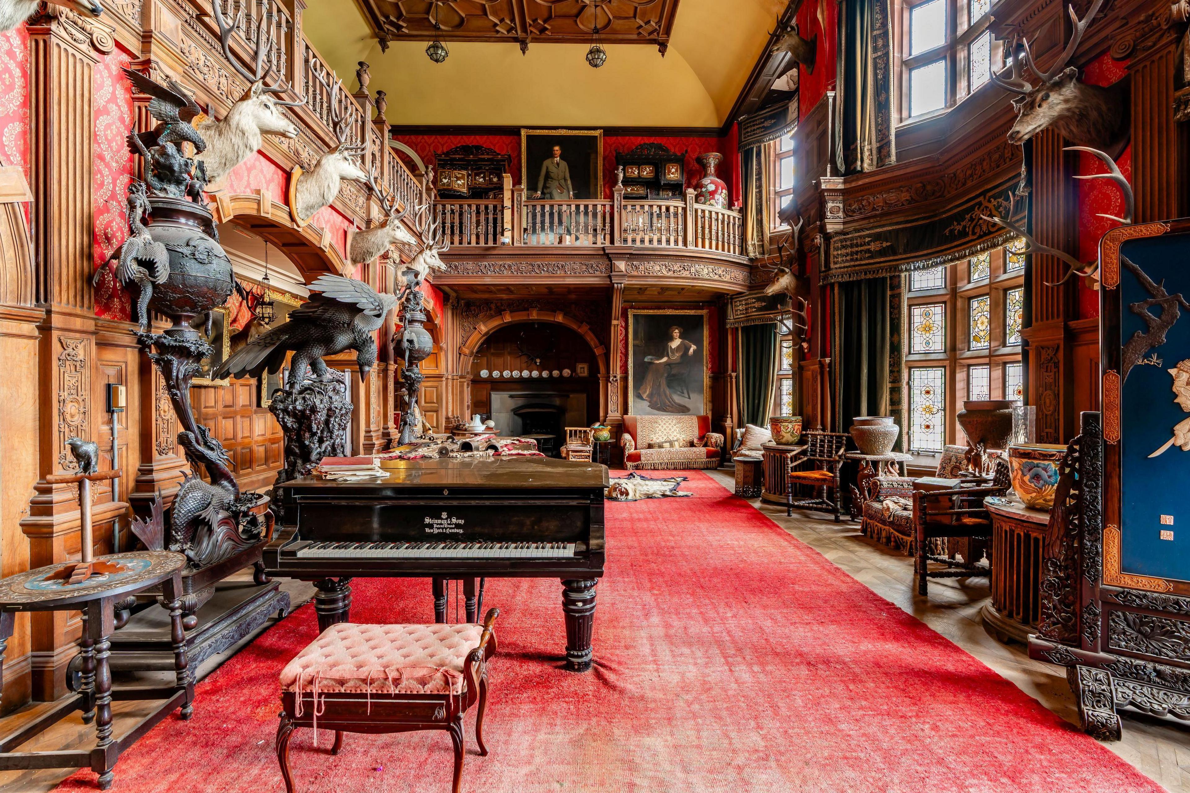 The image shows a large room inside the castle. There is a deep red carpet on the floor, a piano and stags' heads on a wall.