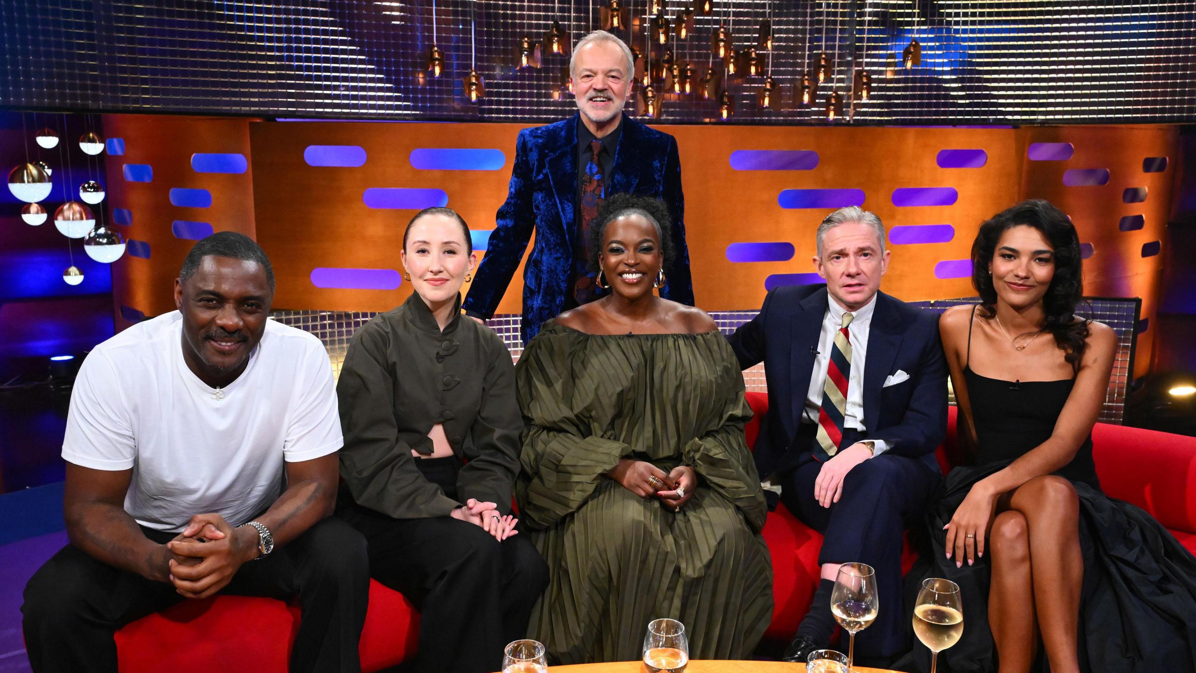 The five Graham Norton Show guests sat on the famous red sofa, with a table covered in drinks glasses in front of them. Behind them stands Graham Norton in a suit with grey hair. Behind the whole group is a brightly-coloured television studio.