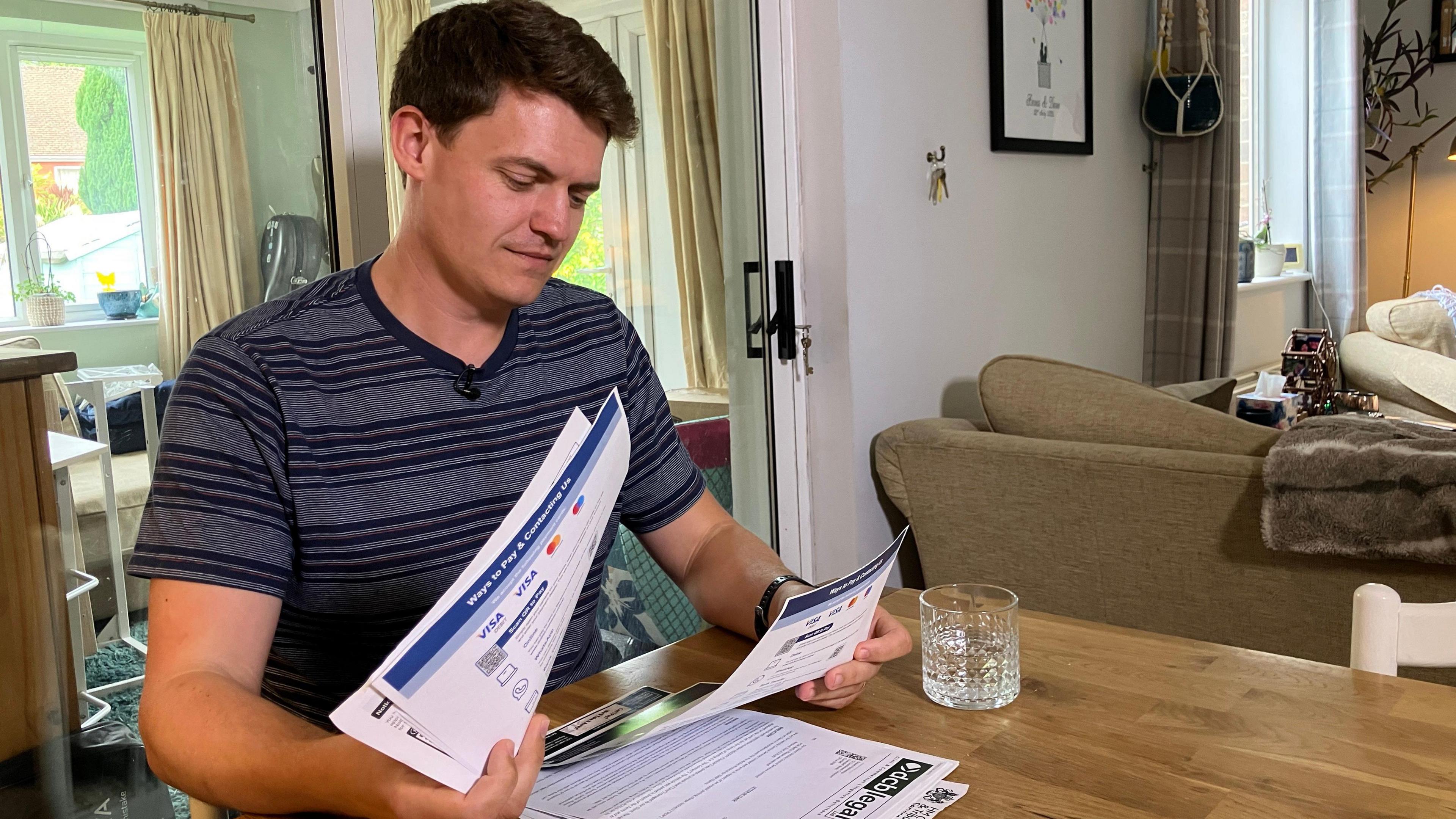 Dave in his living room sitting at a dining table looking through all of the letters he has received. He has short dark hair and is wearing a blue striped t-shirt.