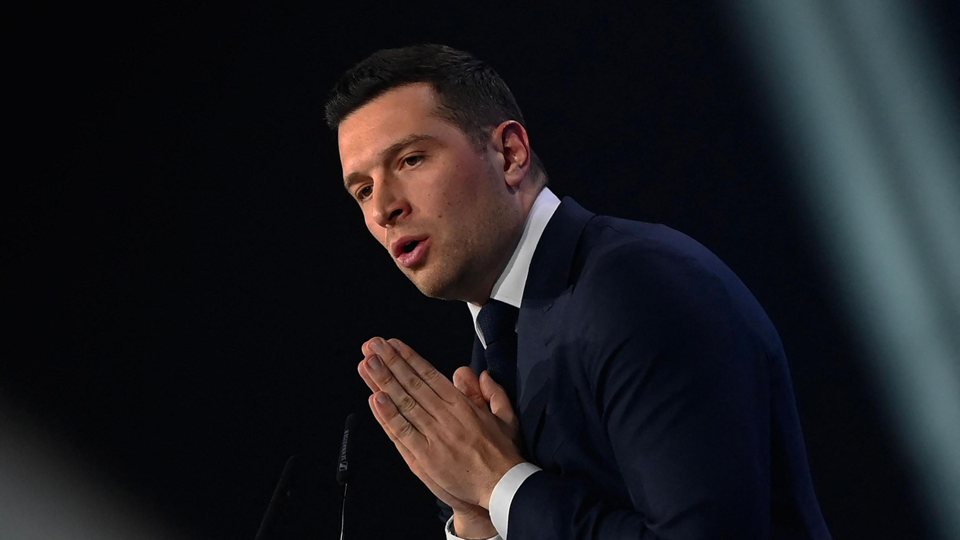 Jordan Bardella wears a suit and stands in front of a dark background with his palms pressed together. 