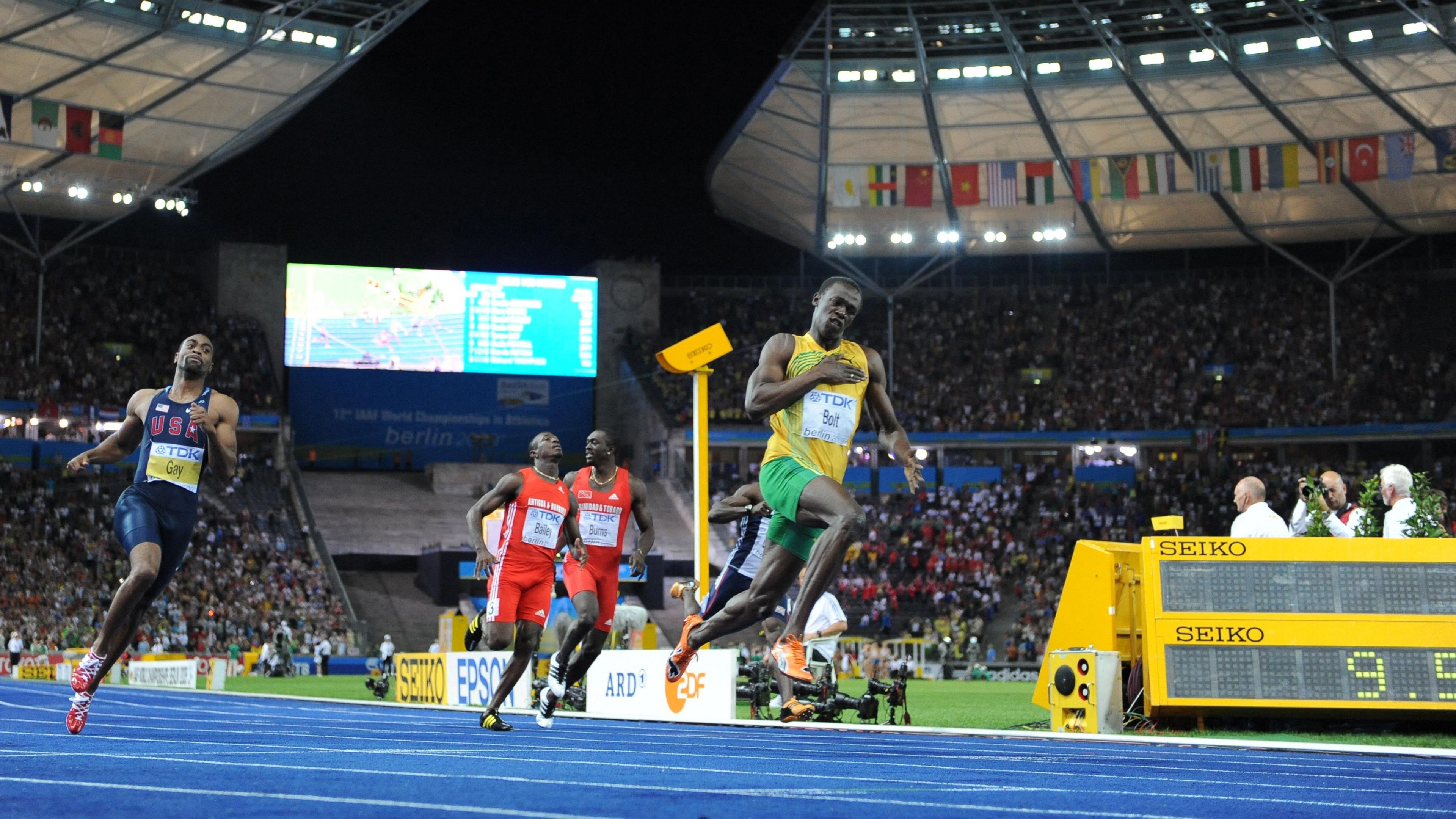 Usain Bolt mid-sprint at the World Athletics Championships in Berlin in Germany in 2009. He's running along a blue track, whilst wearing a yellow vest, green shorts and orange trainers. There are three men running behind him, two wearing red and one wearing navy. They're in a stadium filled with a large crowd.