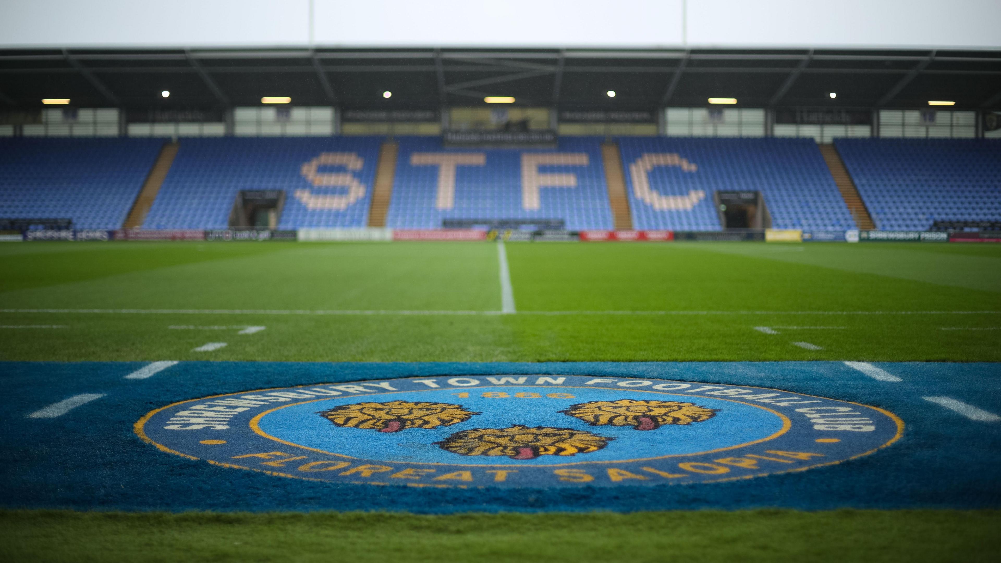 A general view across the pitch at Croud Meadow with the Shrewsbury Town club crest on the grass in the foreground and the seats in the stand opposite showing the letters STFC