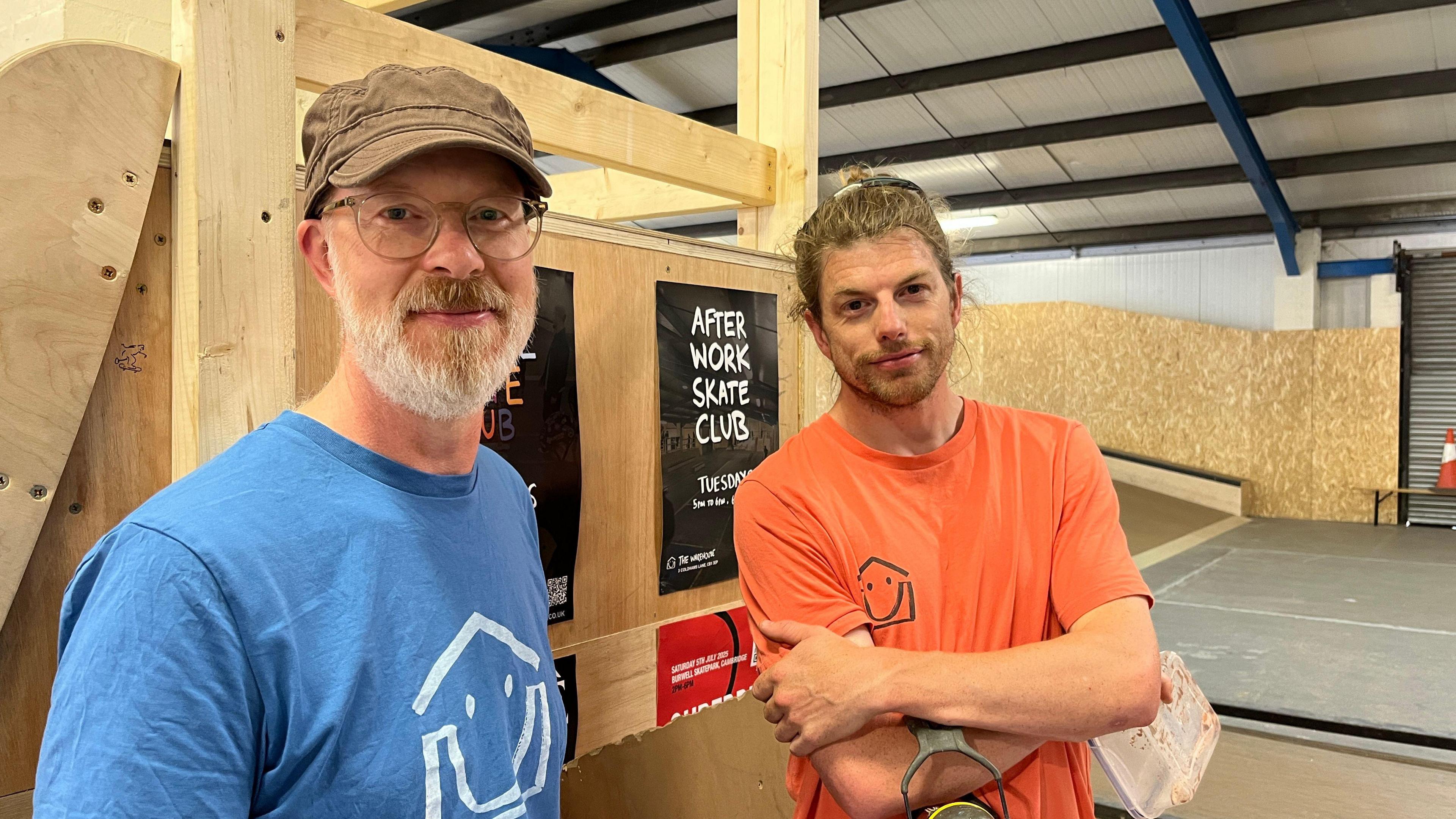 Two members of Cam Skate are standing at the site, which is made up of wooden beams and ramps inside a warehouse. The two men are smiling and looking at the camera. Nearest the camera, a man is in a khaki hat, has a white beard and a blue T-shirt. In the background, the man has long hair tied back and has an orange T-shirt on.