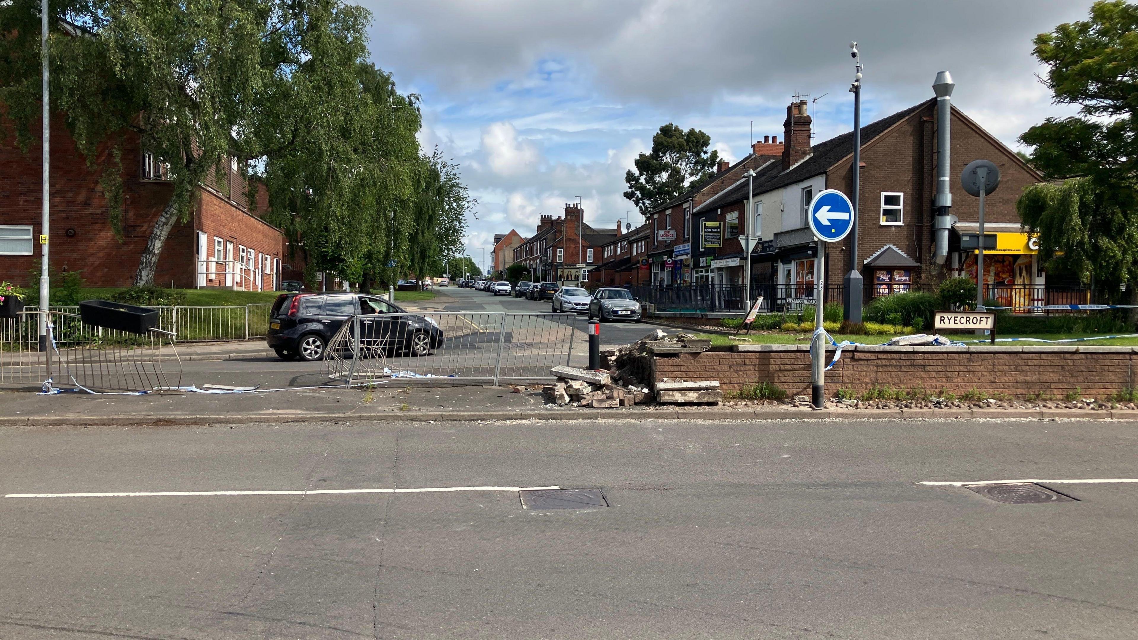 A street scene shows a damaged brick wall and temporary fencing, suggesting a recent crash. Cars and shops line the road under a cloudy sky.

