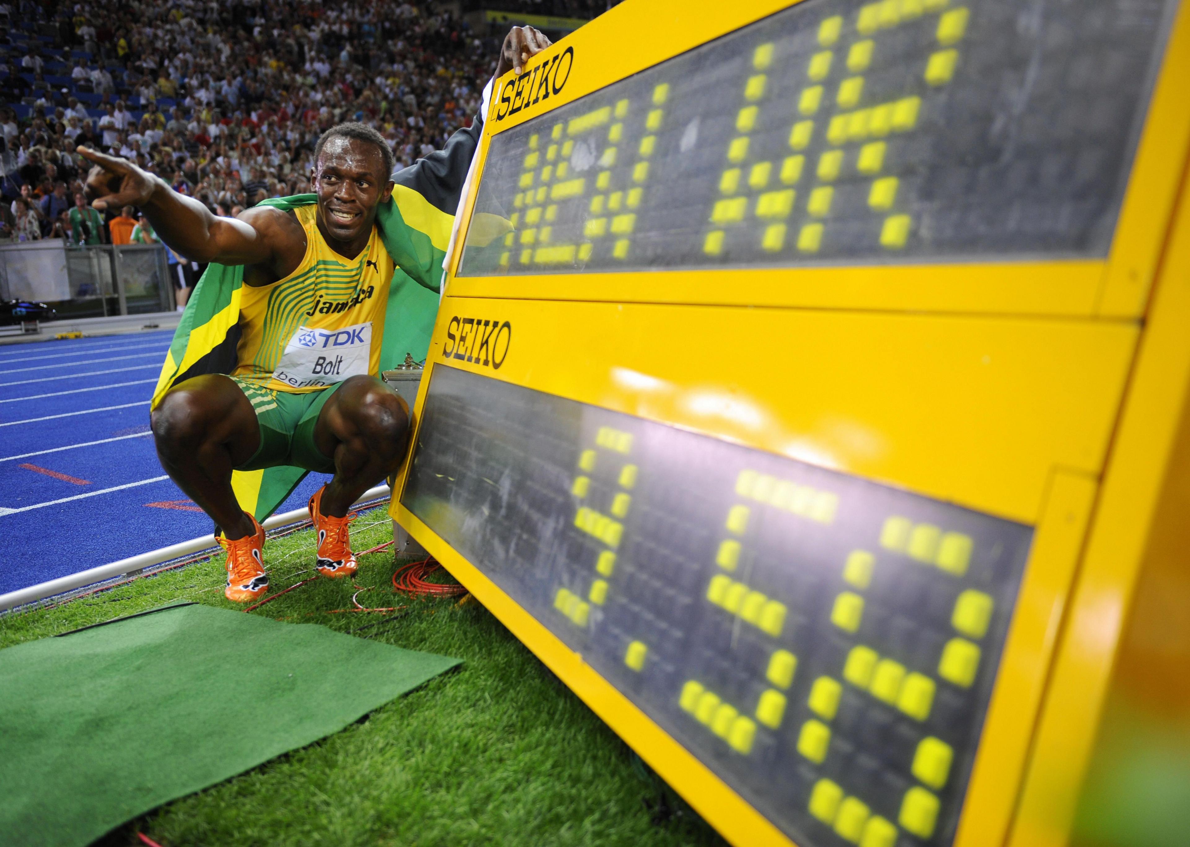 Usain Bolt crouches in front of an electronic sign saying "New WR" with the time 9.58. He is by the side of an athletic track, pointing into the distance with the Jamaican flag wrapped around his shoulders.