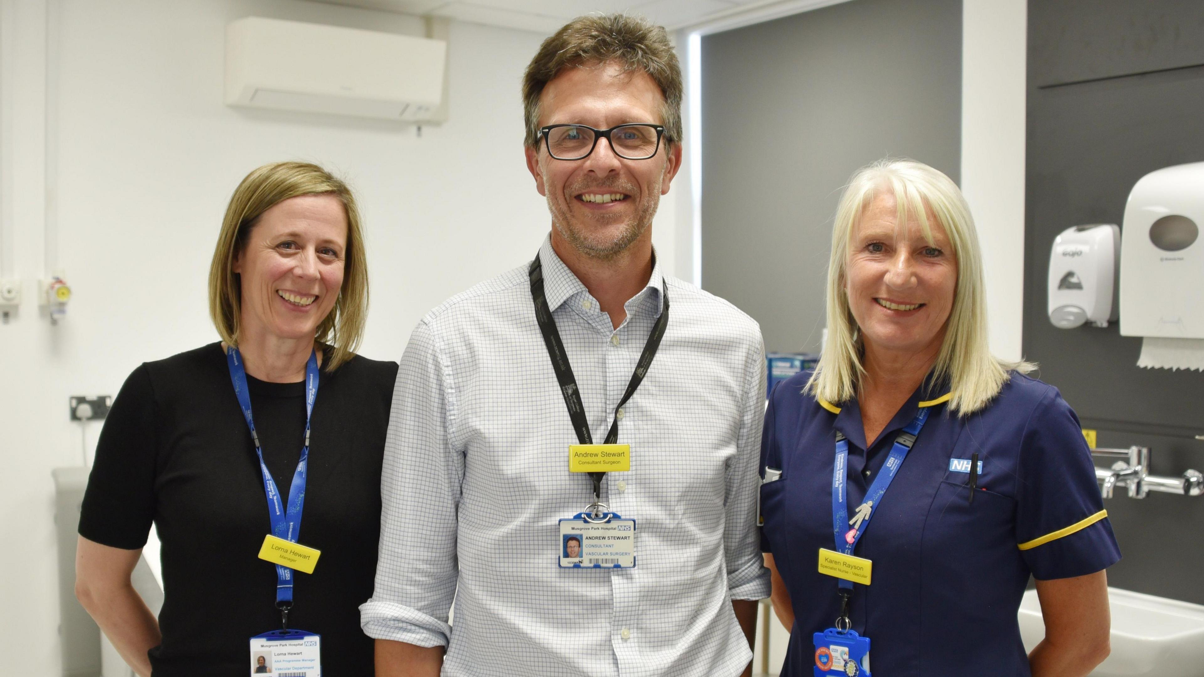 Two female doctors wearing blue lanyards and their hospital scrubs. They both have shoulder length blonde hair and are smiling at the camera. They are standing either side of a tall male doctor in the middle, who is wearing a white checked shirt and a black lanyard. They are all standing in the new treatment room.