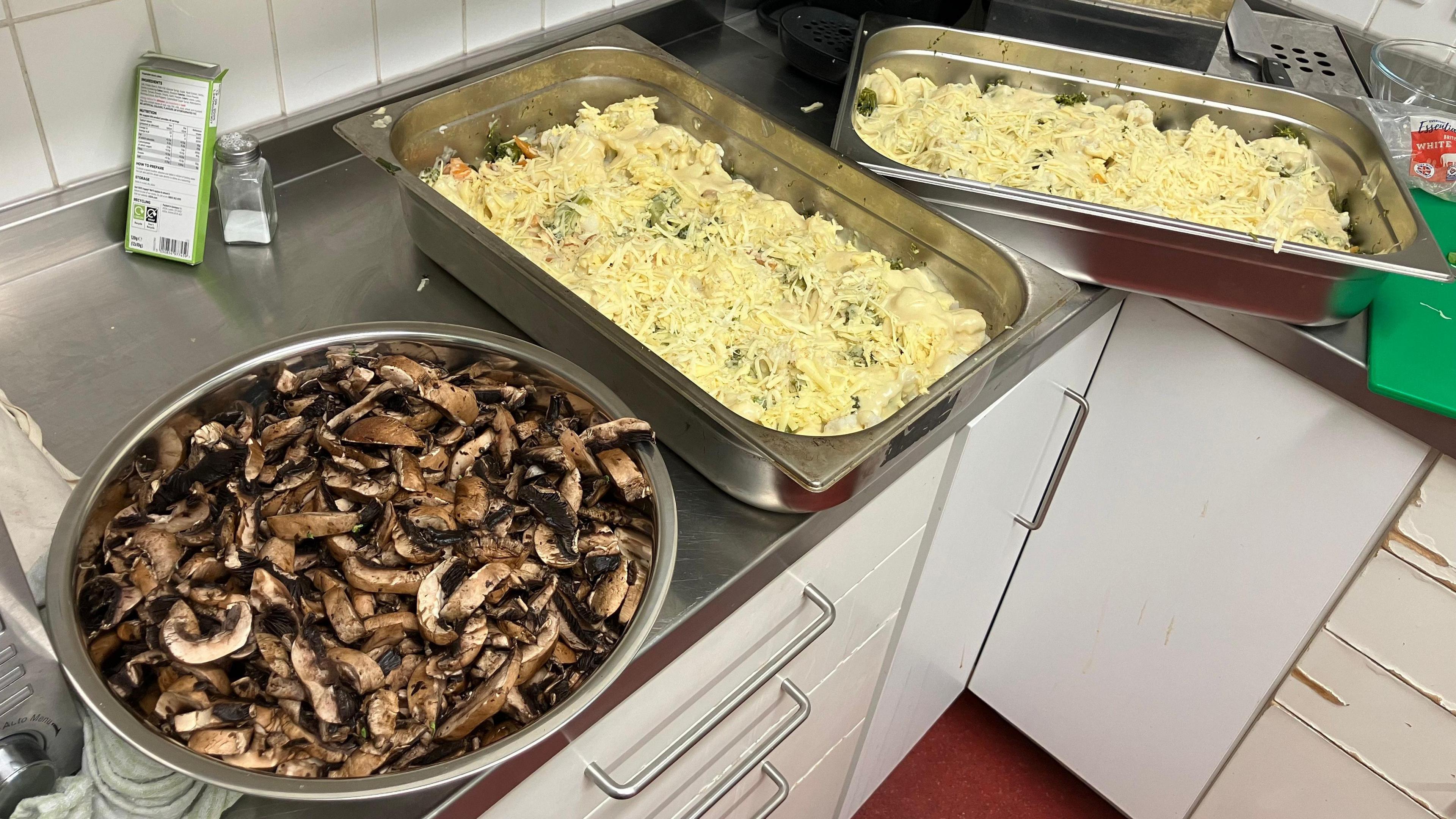 A large silver bowl with sliced mushrooms and a large rectangular serving  dish with cheese and creamy food inside. The containers are on a stainless steel work surface in the community centre kitchen.