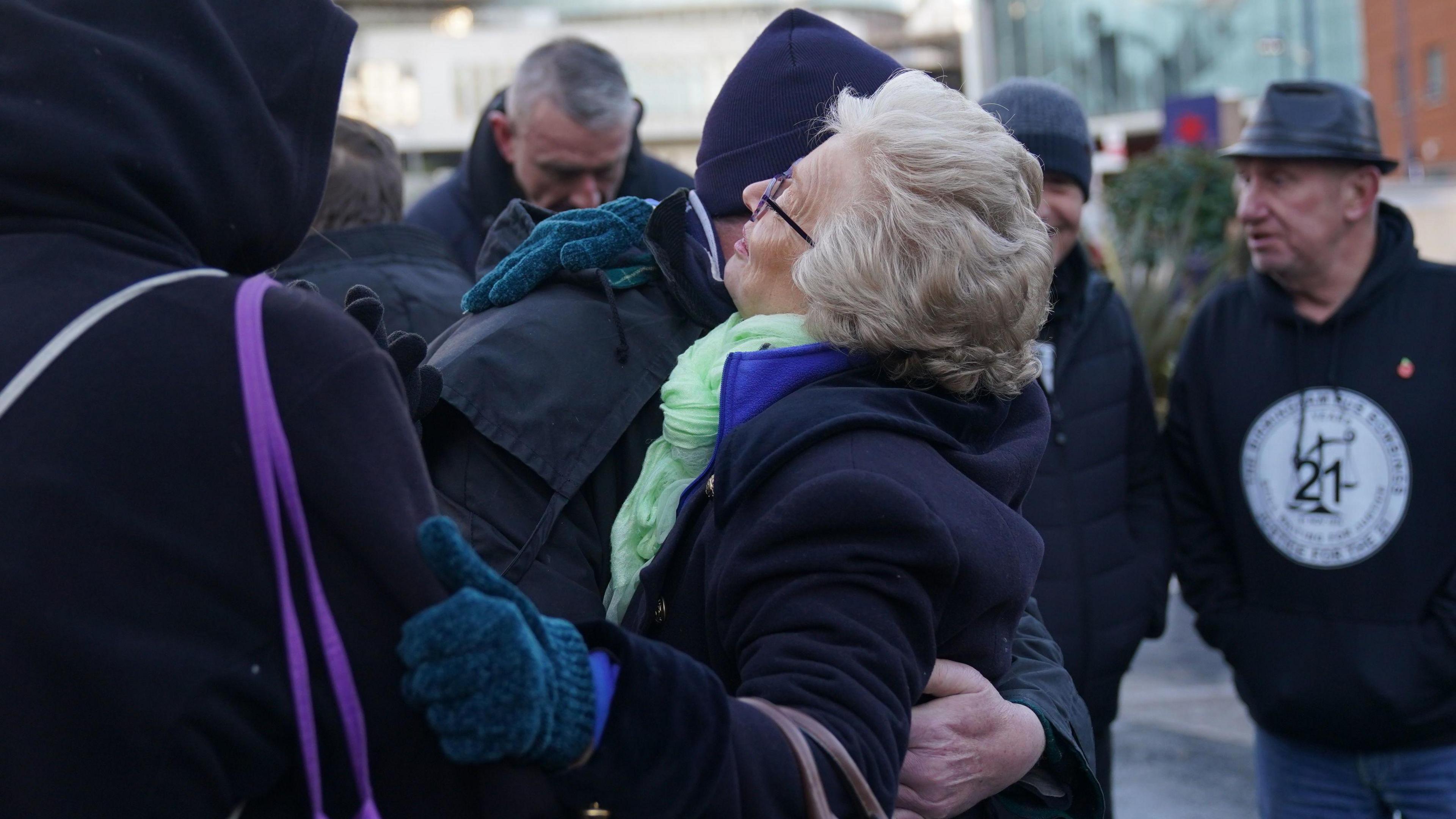 Julie Hambleton is seen hugging a fellow campaigner at a memorial service. She is wearing a coat and scarf and gloves and people around her, outdoors in the city centre, are also in hats and coats. One man has his head bowed.