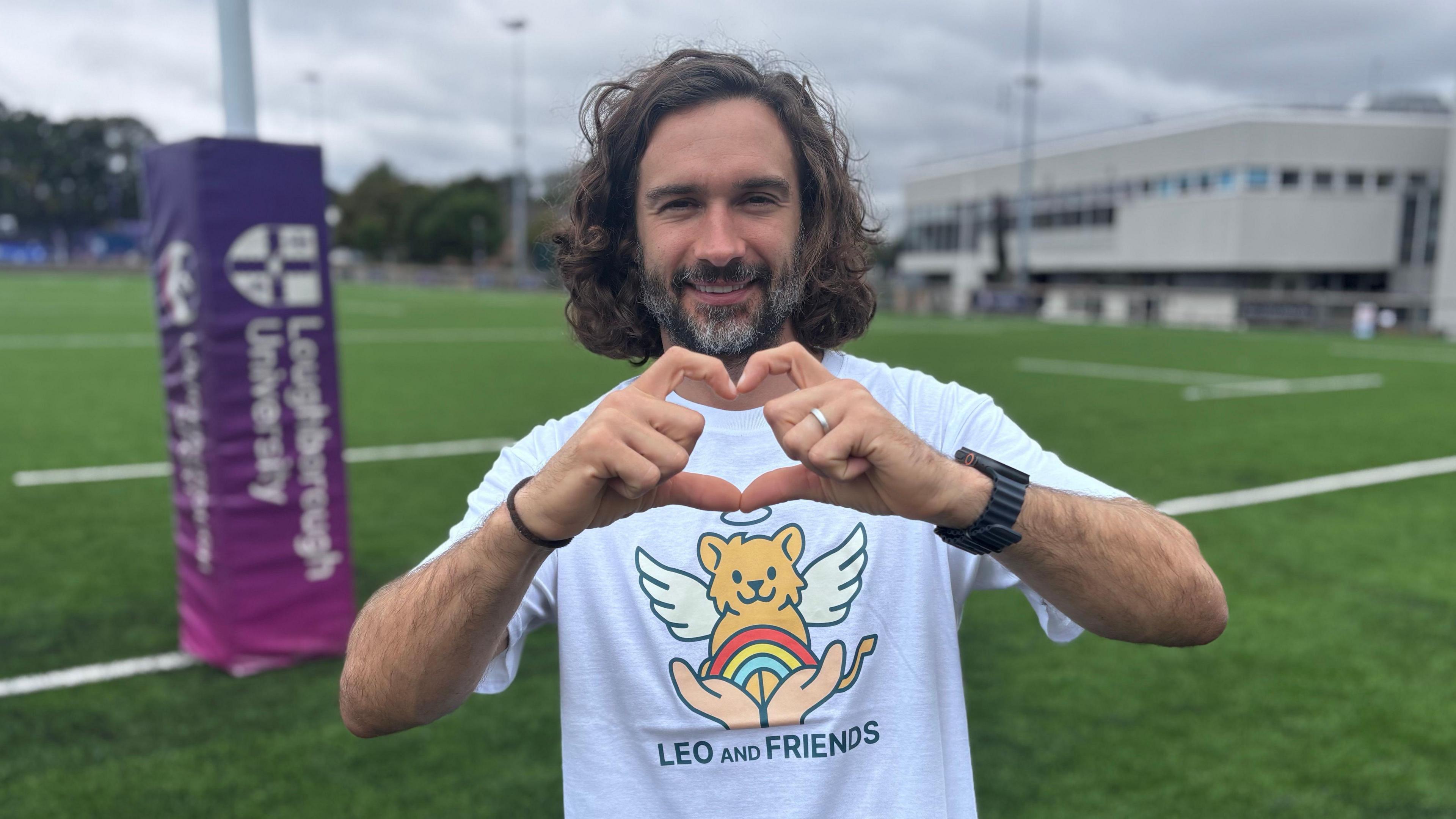 Fitness coach Joe Wicks wearing a white t-shirt with a Leo and Friends motif. He is standing on a Loughborough University sports pitch and smiling, while making a heart shape with his hands.