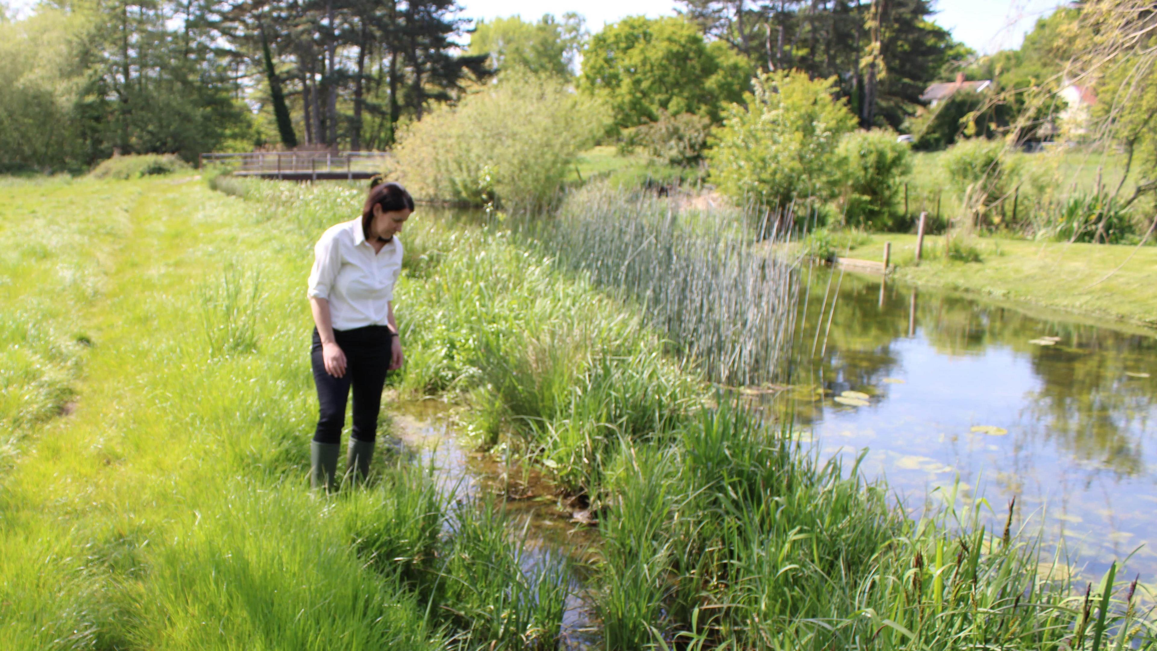 Dr Helen Dangerfield walks on the edge of the River Deben during a sunny day. She wears a white shirt with black trousers and wellington boots. She has dark hair that is partly tied back.