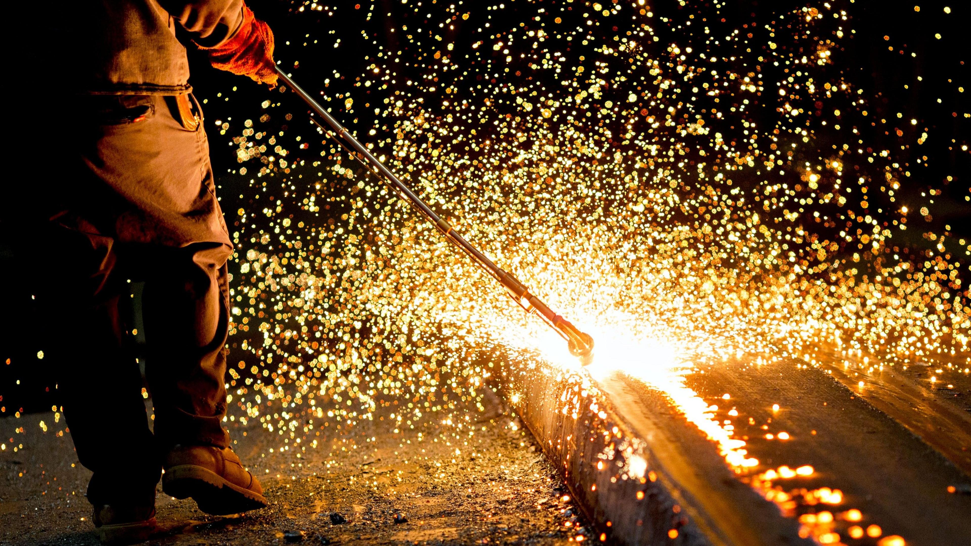 A steelworker runs a long metal tool along a steel plate causing countless sparks to fly in all directions.