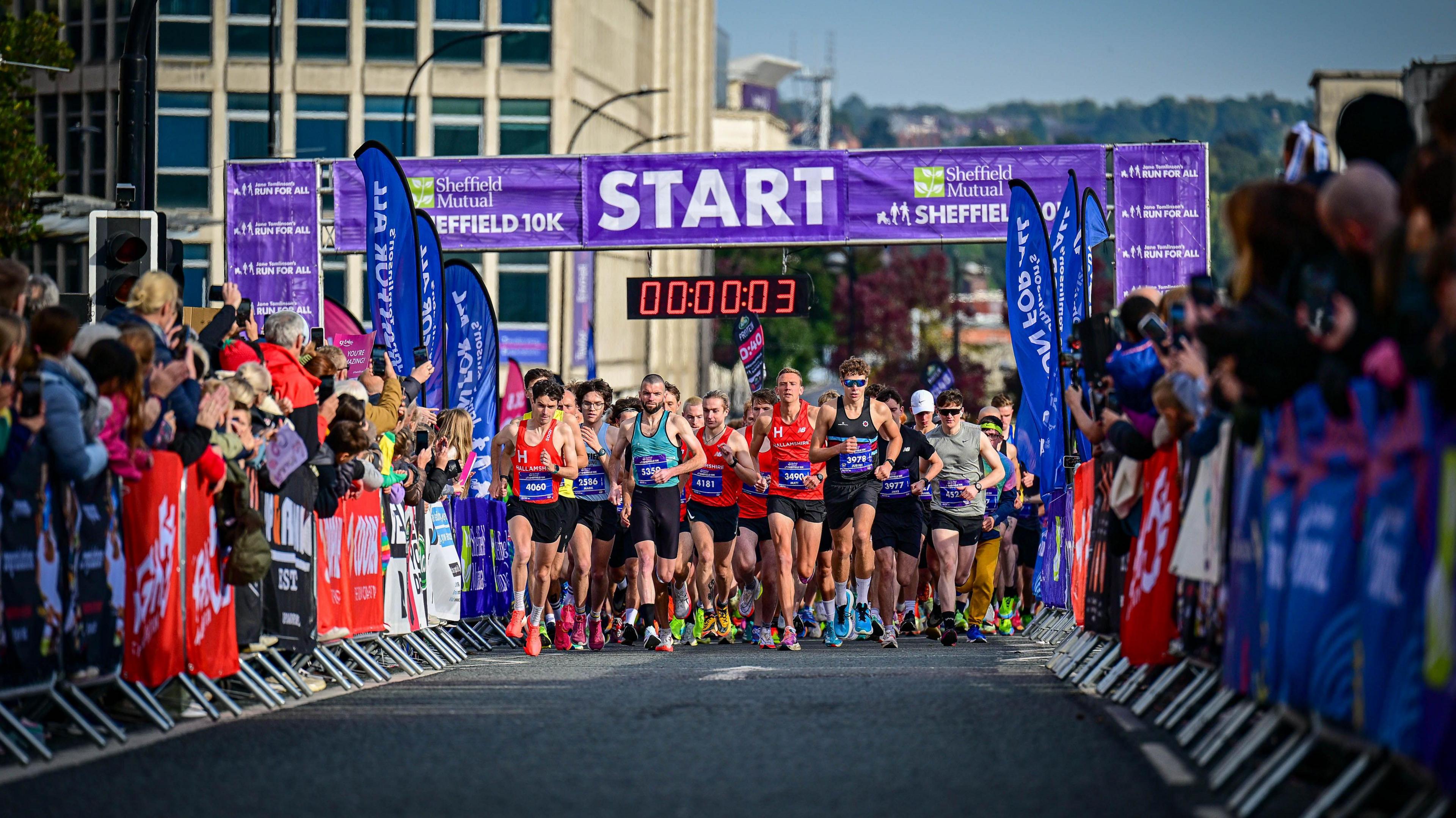 Runners line up beneath a large blue 'start' banner covered in Sheffield 10k branding . Along each side of the road are metal barriers lined with spectators