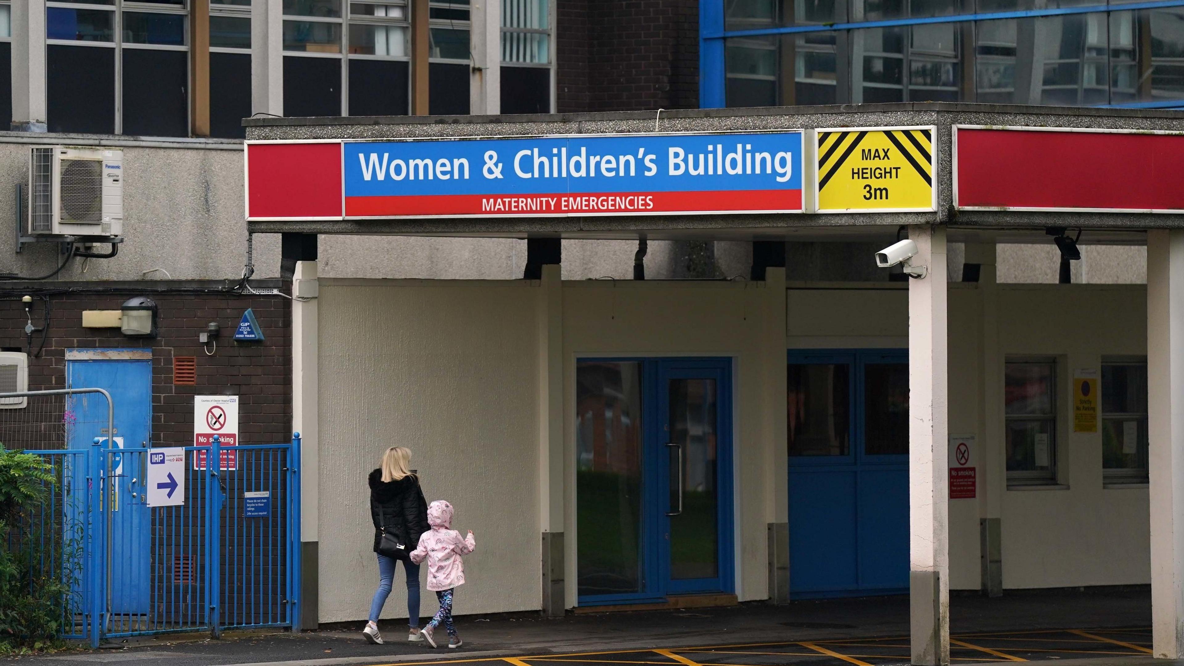 A woman and her young daughter, wearing a pink coat, outside a hospital building walking towards a set of blue doors under a sign reading "Women & Children's Building - Maternity Emergencies". 