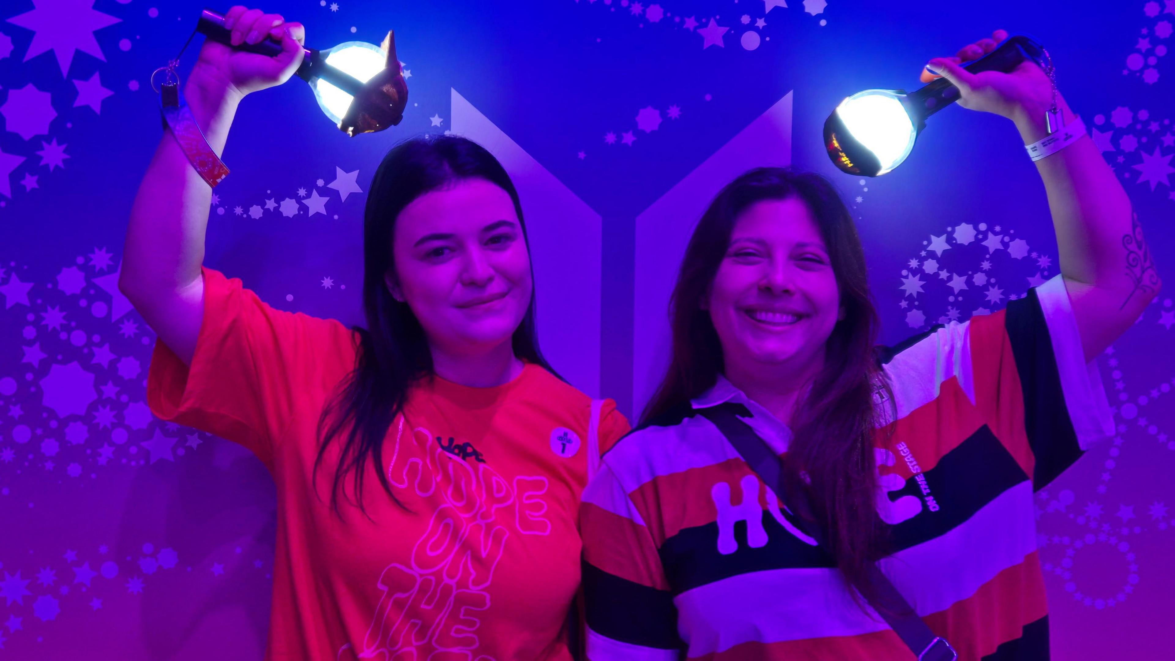 Stephanie Prado, left, wearing a red shirt, poses with her friend while holding an official BTS light stick in her right hand