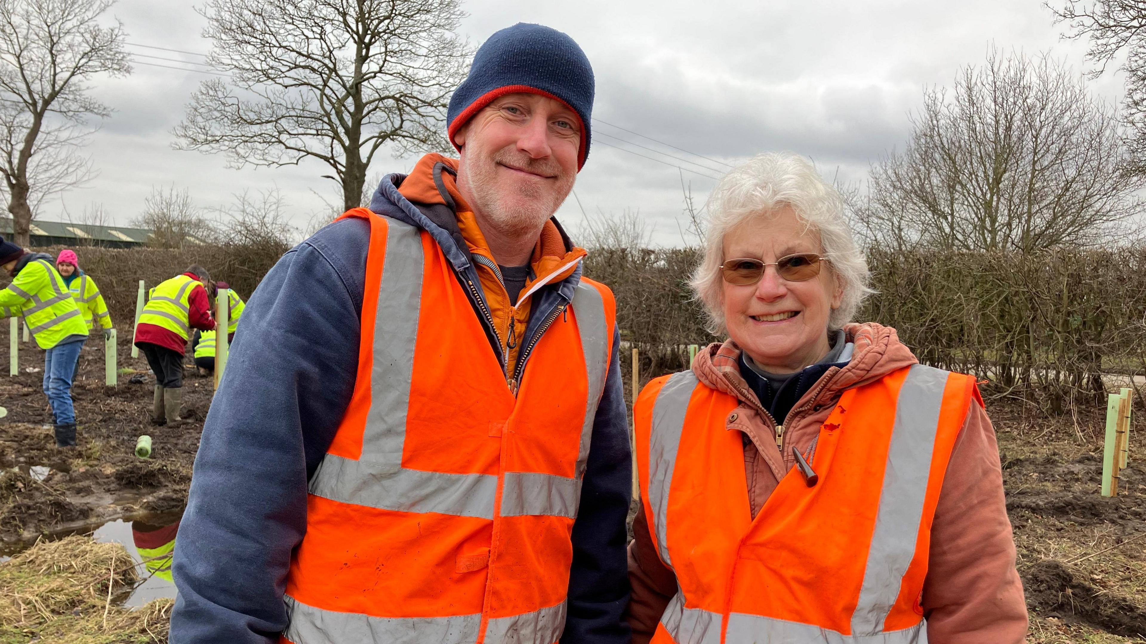 A man and a woman stand smiling together, with both wearing reflective vests. The man is wearing a blue hat. The older woman has white hair and glasses. In the background, volunteers are planting trees.