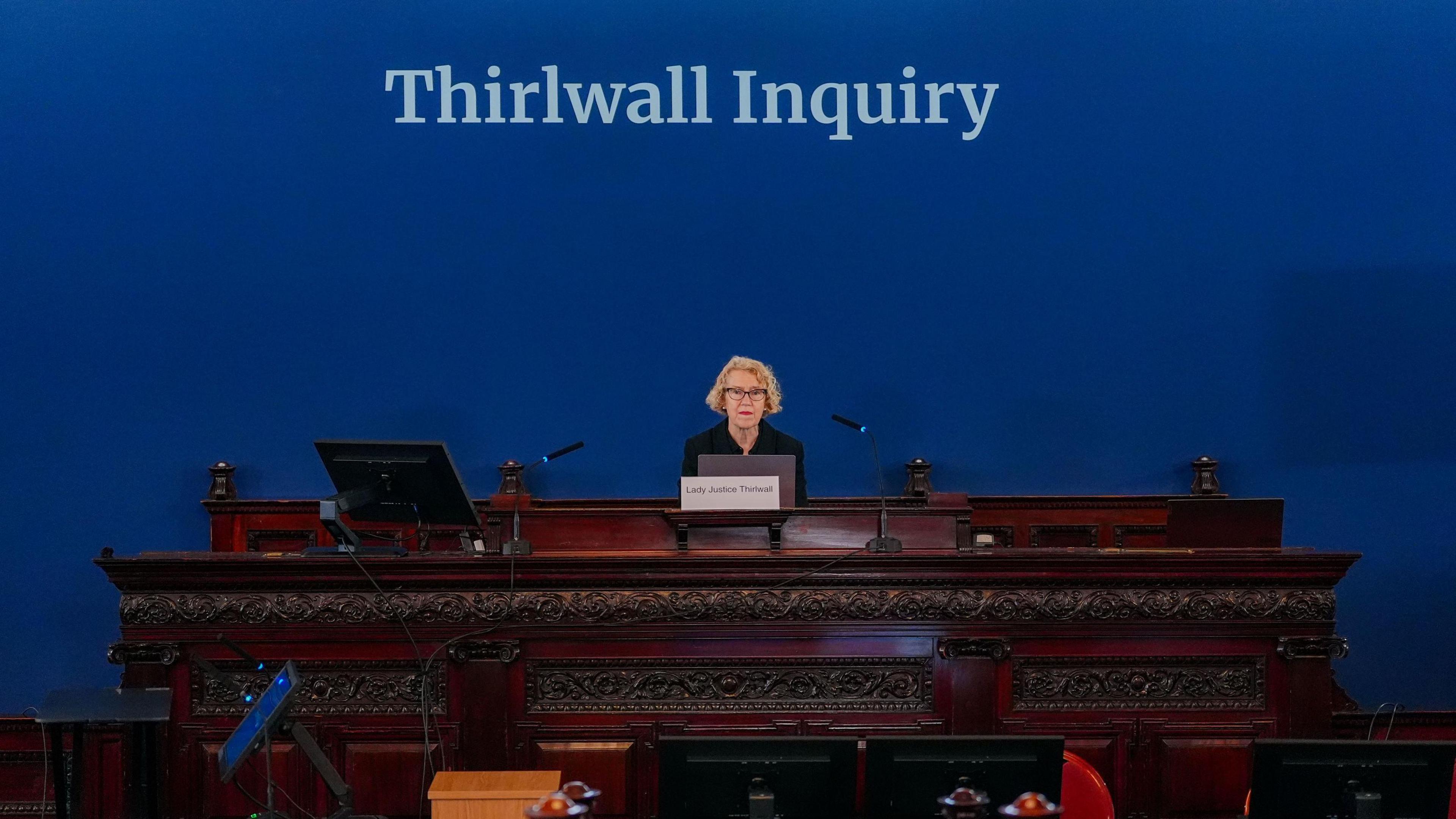 Chair of the inquiry Lady Justice Thirlwall at Liverpool Town Hall. she has blonde curly hair and sits on a burgundy wood pew in front of a blue background with Thirlwall Inquiry written above her.