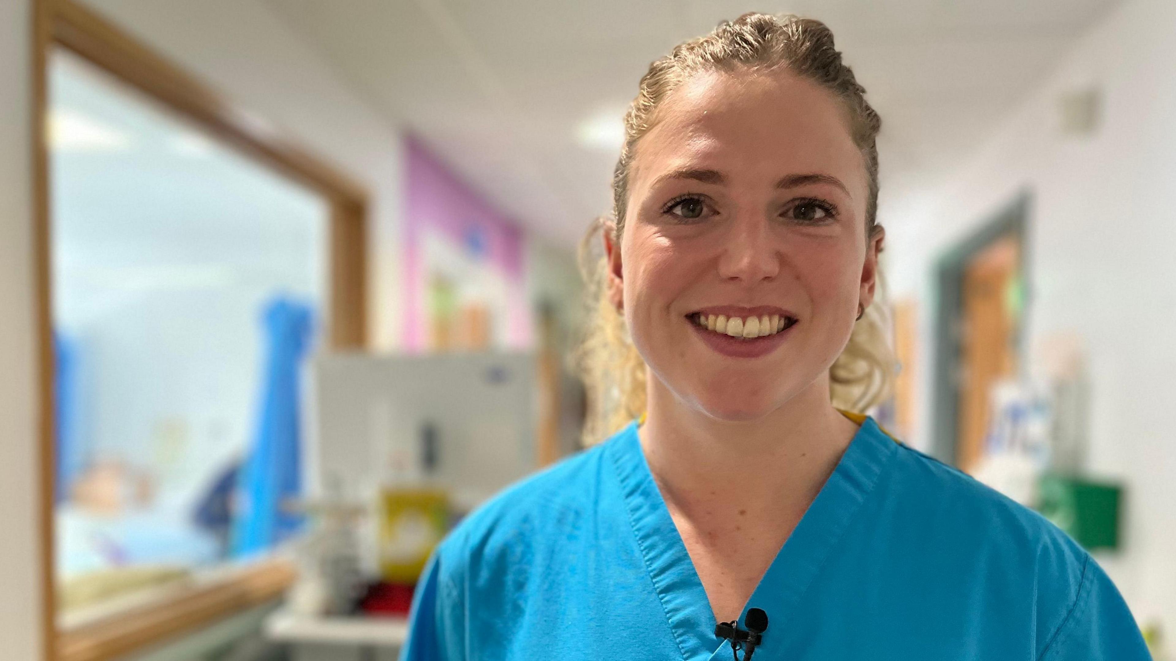 Kerenza Bryson is looking directly at the camera and smiling. Her hair is blonde and curly and pulled back into a ponytail. She is wearing blue medical scrubs. 