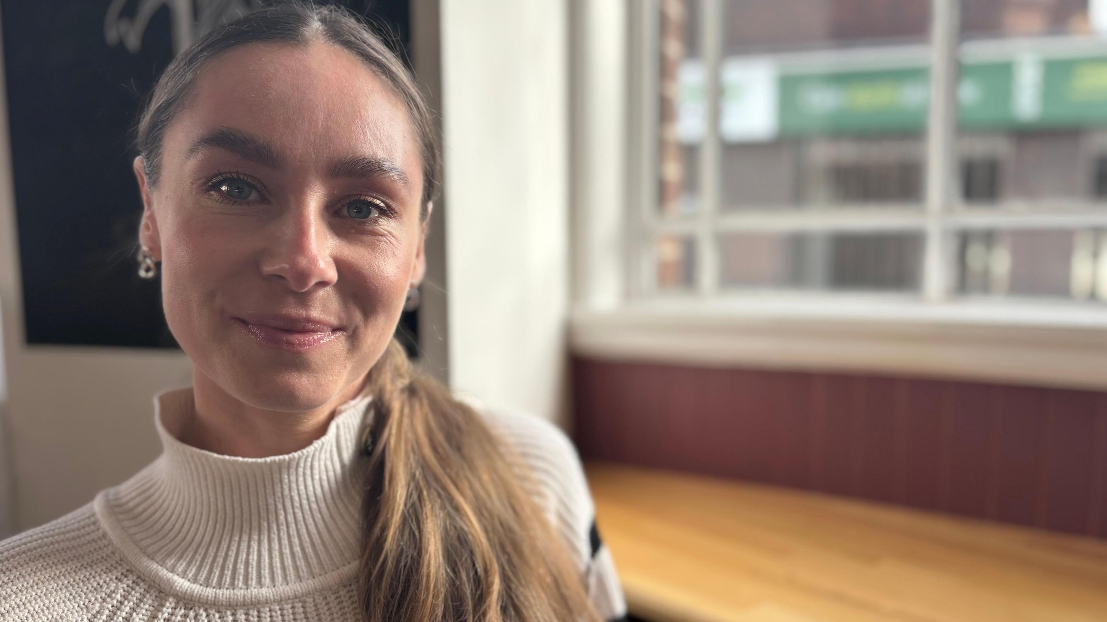 Natalie with long light brown hair in a pony tail wearing a high neck cream jumper sitting in a cafe by a window. She is smiling.