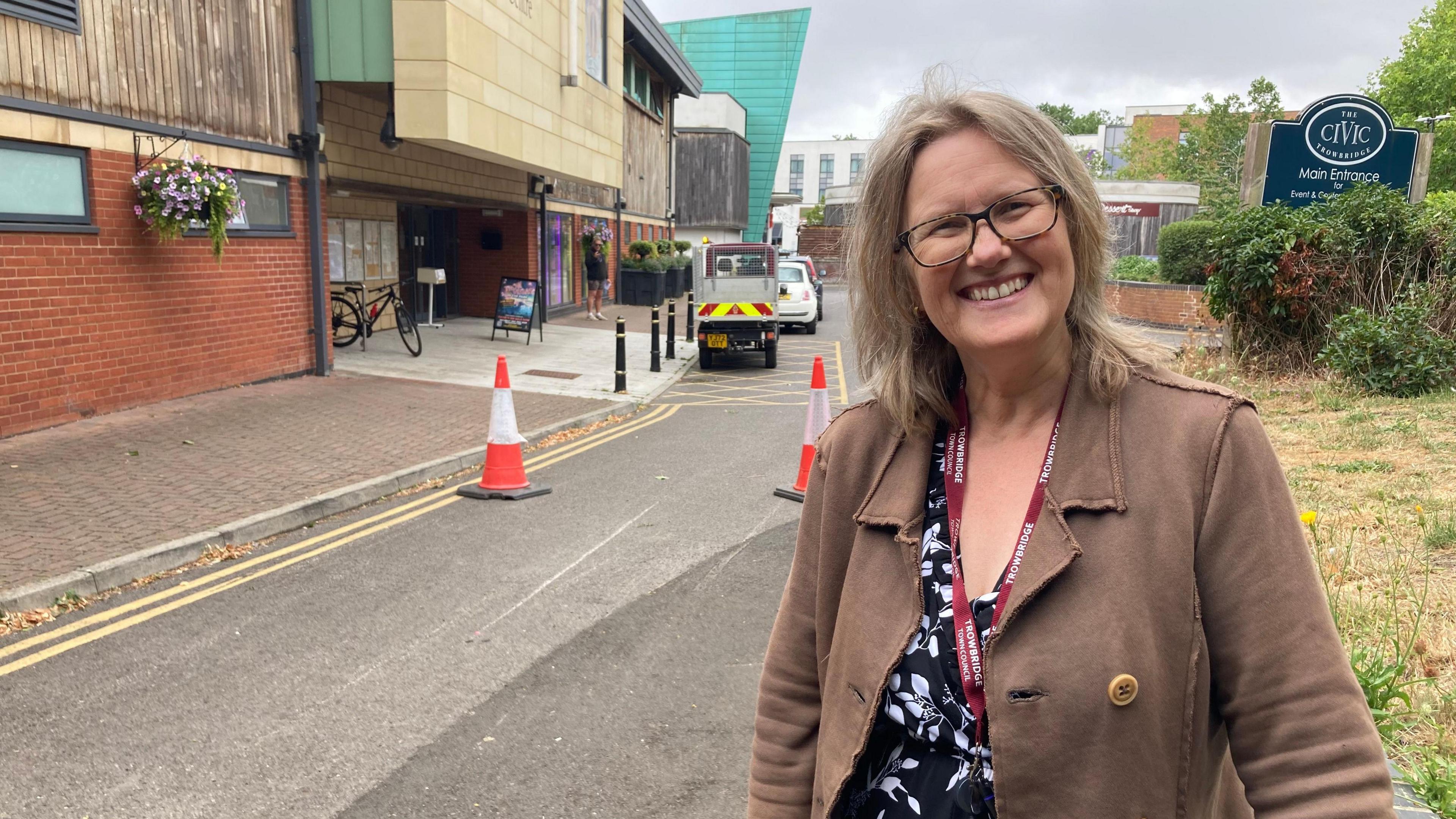 A middle-aged woman with lightly coloured hair and glasses smiles at the camera. She's wearing a brown jacket, a lanyard and a black and white shirt standing in front of a building and sign that reads "The Civic Trowbridge".