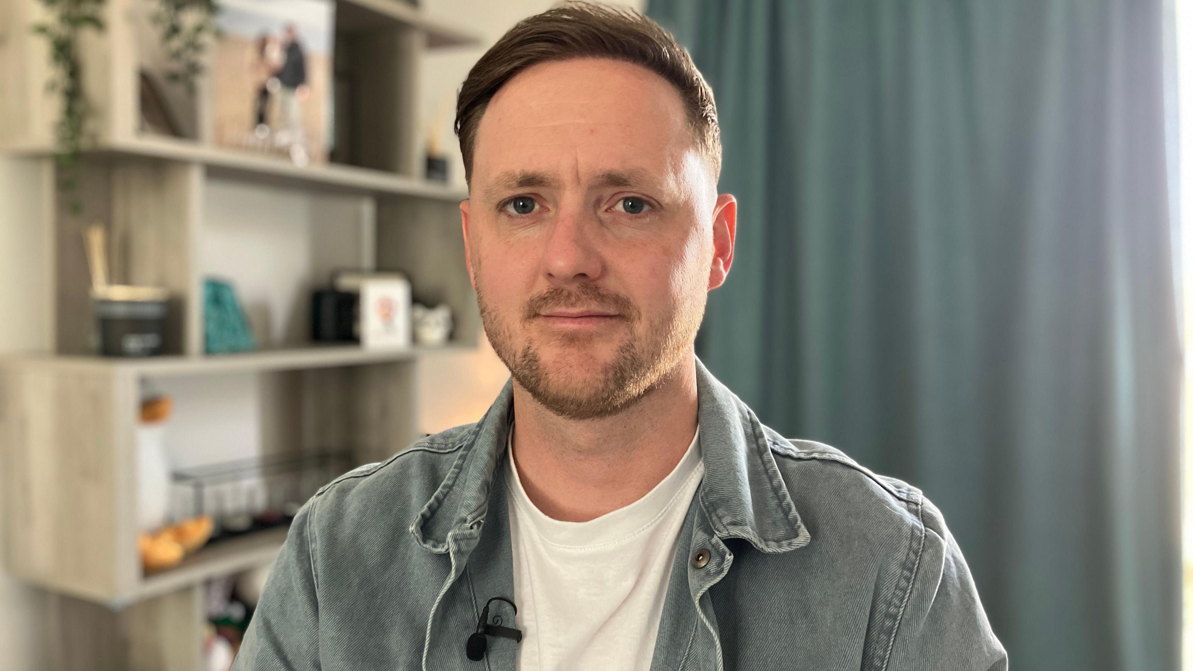 Chris Rodgers. He has short brown hair and facial hair. He is looking directly at the camera and smiling. He is sat inside a house with shelving, photo frames and a curtain in the background.
