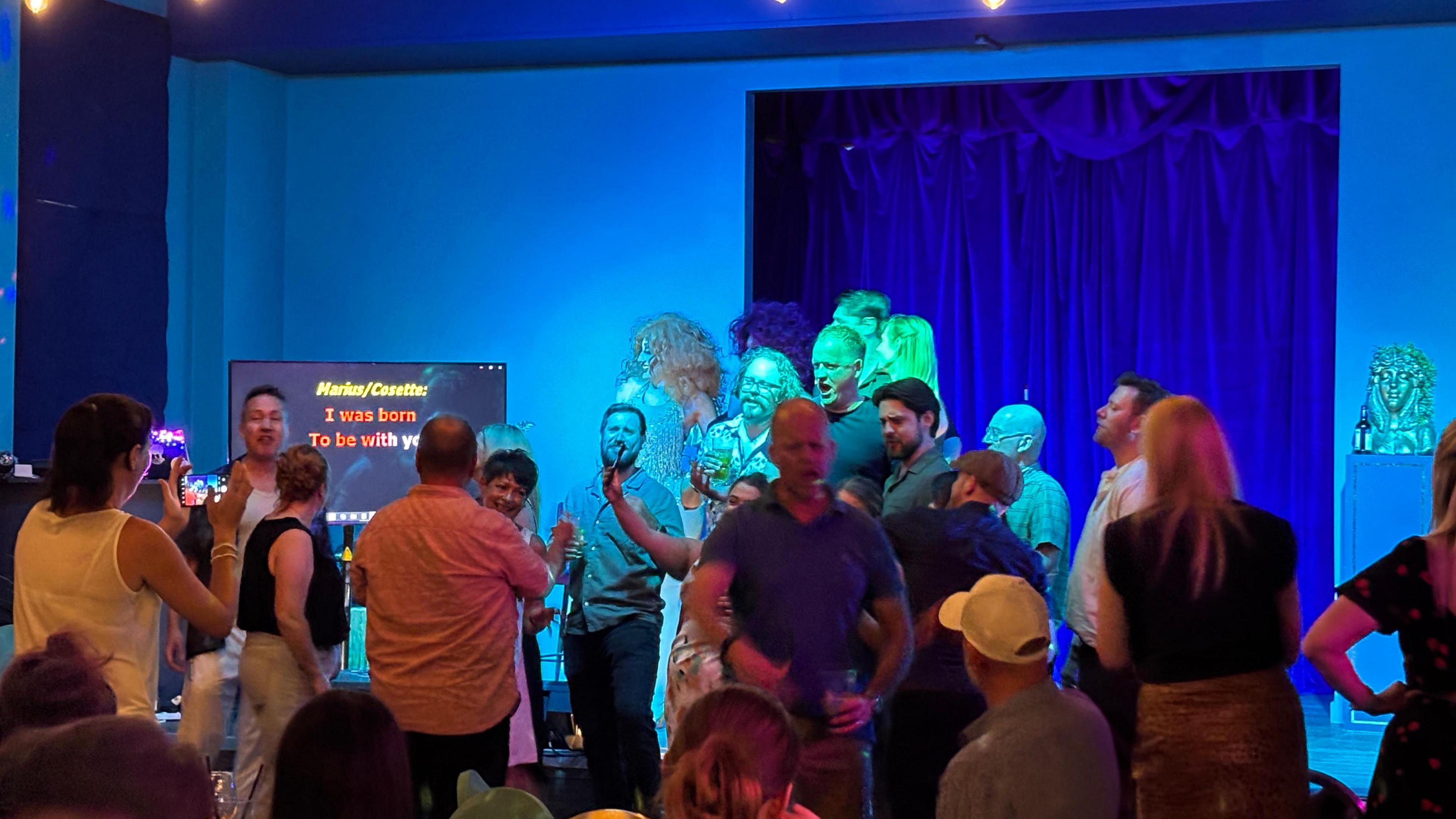 People partying in a hall and singing along to karaoke. It is dimly lit.