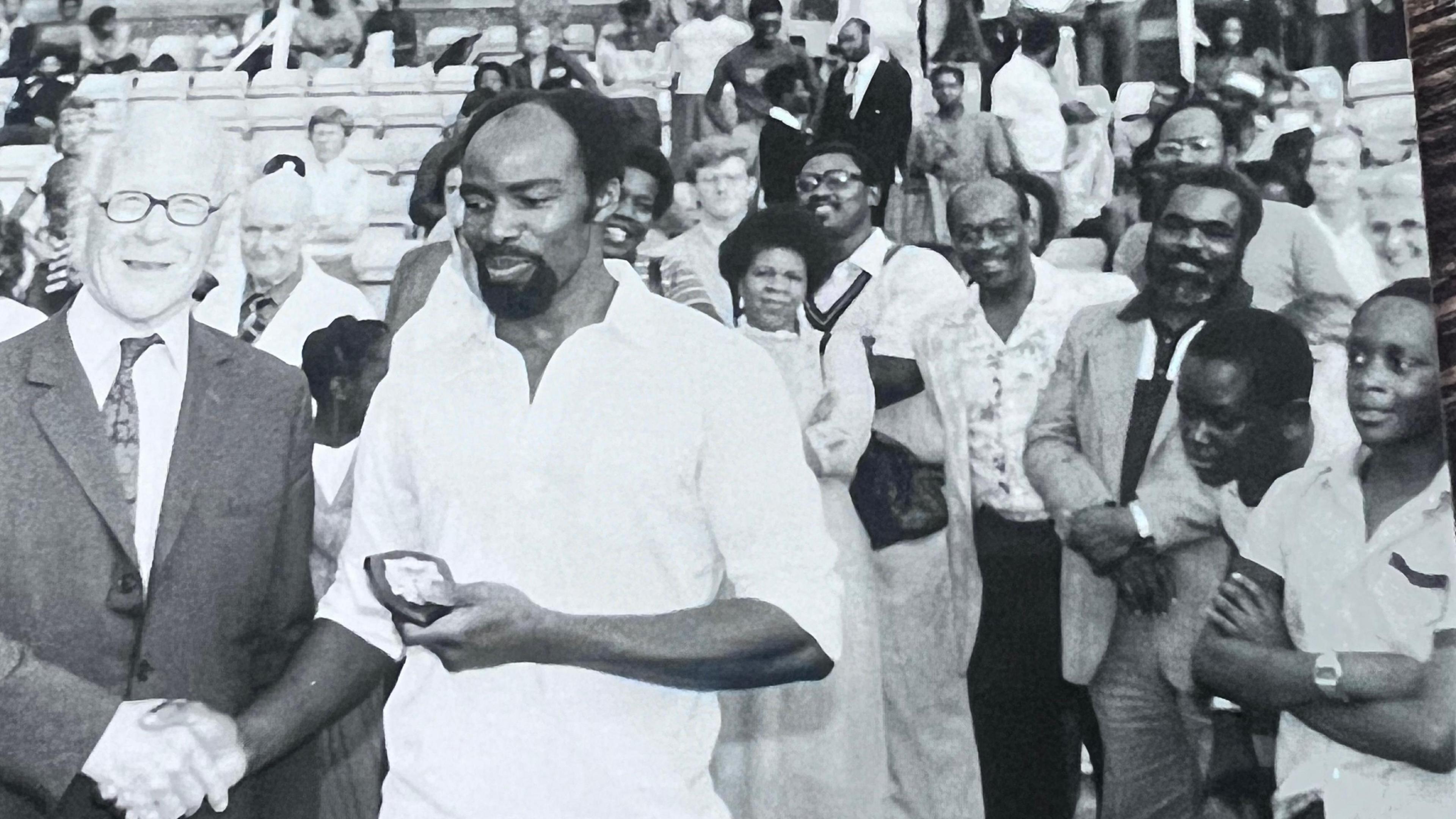 A black and white image from 1982, showing cricket coach and former Brixton West Indies player and captain Neville Walker shaking hands with a man, as he holds a plaque. The photograph is taken at a cricket ground, and spectators can be seen standing directly behind the pair, whilst some can be seen sitting in the background.”