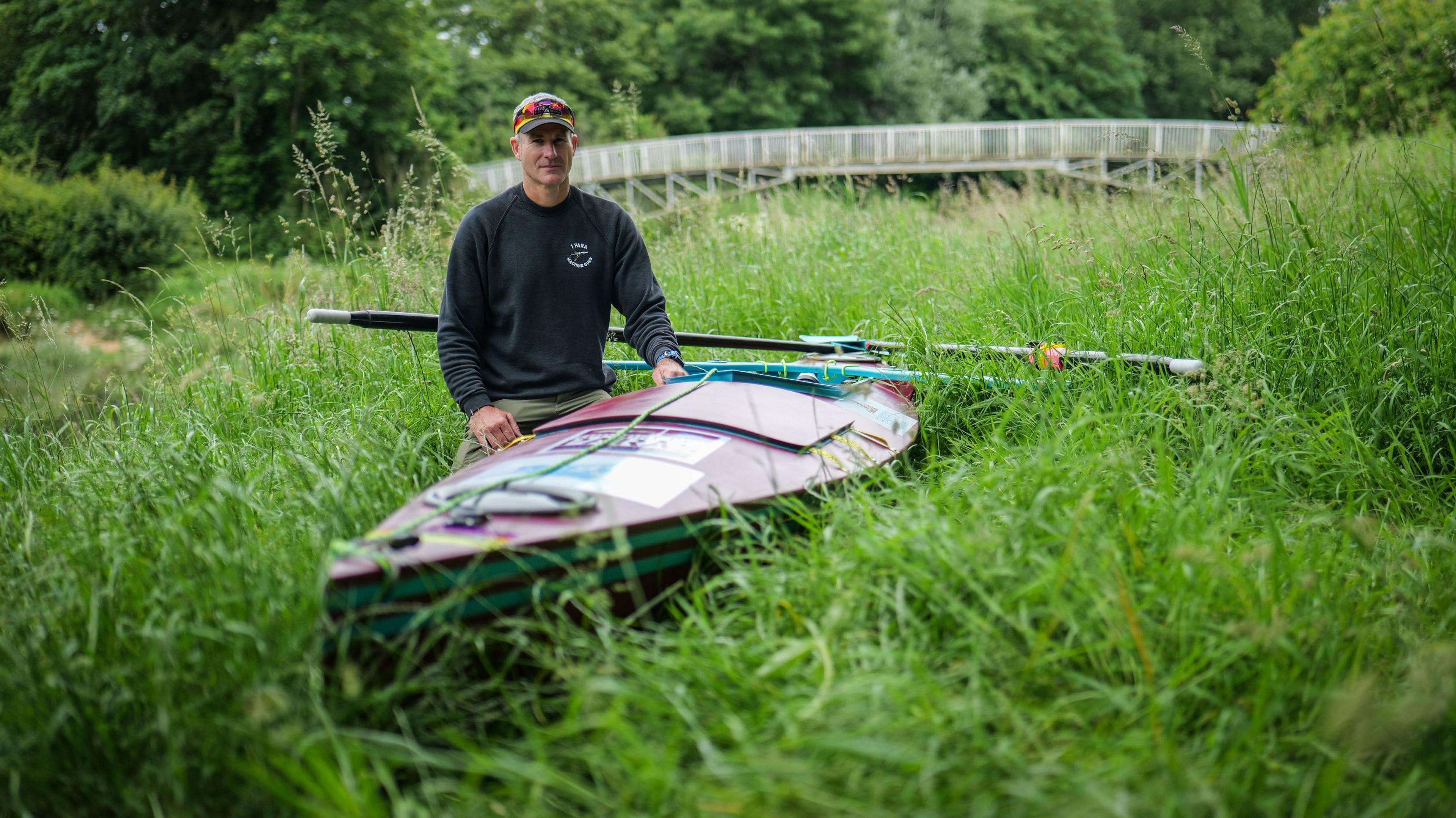 Mike Ellicock, wearing a grey sweatshirt, green trousers, baseball cap and sunglasses, sits beside his canoe on dry land.