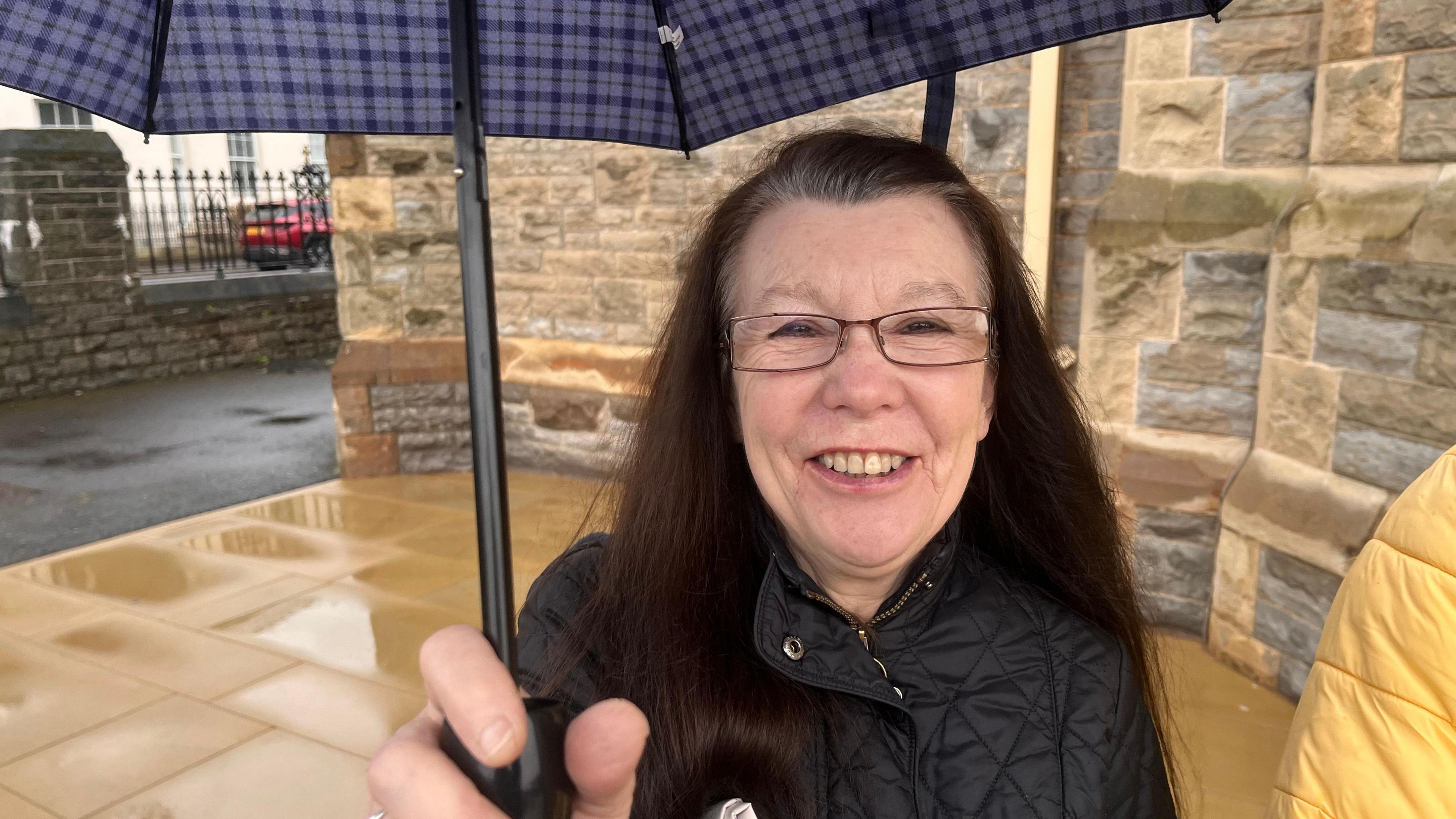 Anne, who has long, dark hair and wears glasses and a black coat and holds an umbrella and she stands outside Clonard Monastery, which is a light-coloured stone building.