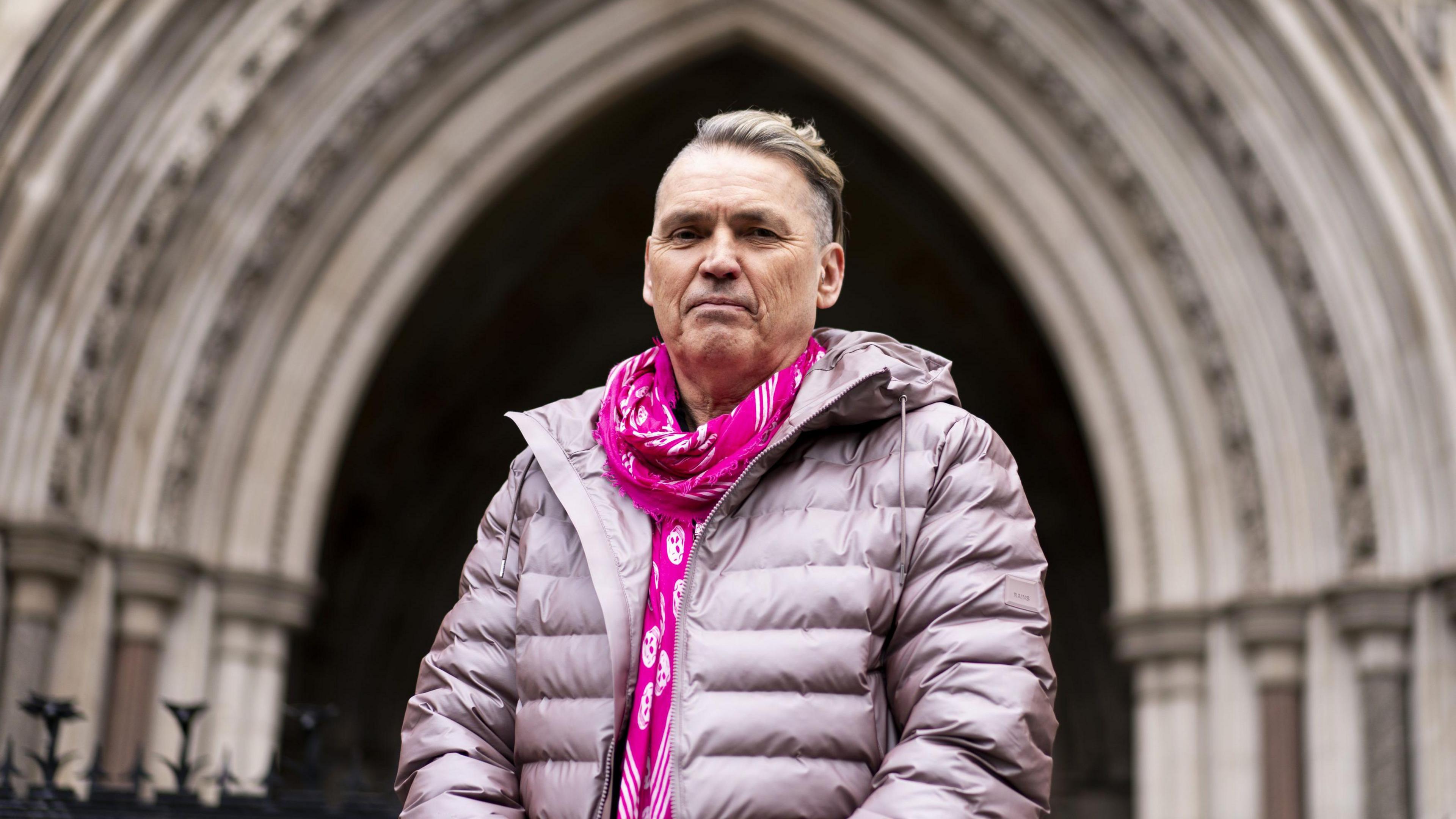 Dale Vince, an older man wearing a purple puffer coat and hot pink scarf with white skulls on, stands outside the entrance to the Royal Courts of Justice in London