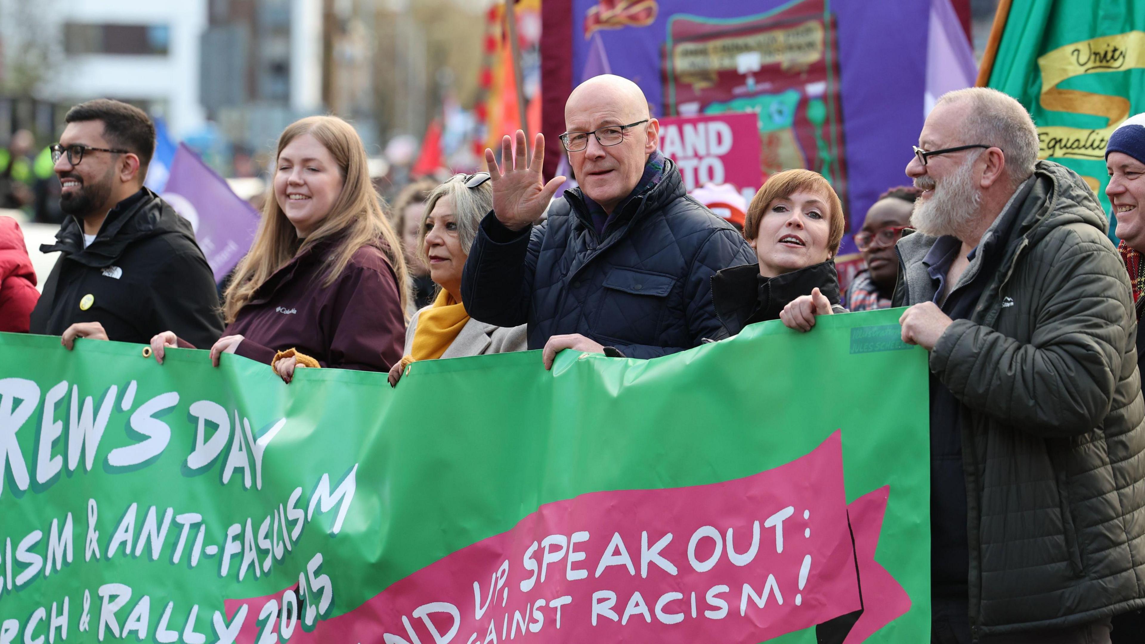 John Swinney at an anti-racism rally - he is one of several people holding a large green banner that reads "stand up, speak out against racism".