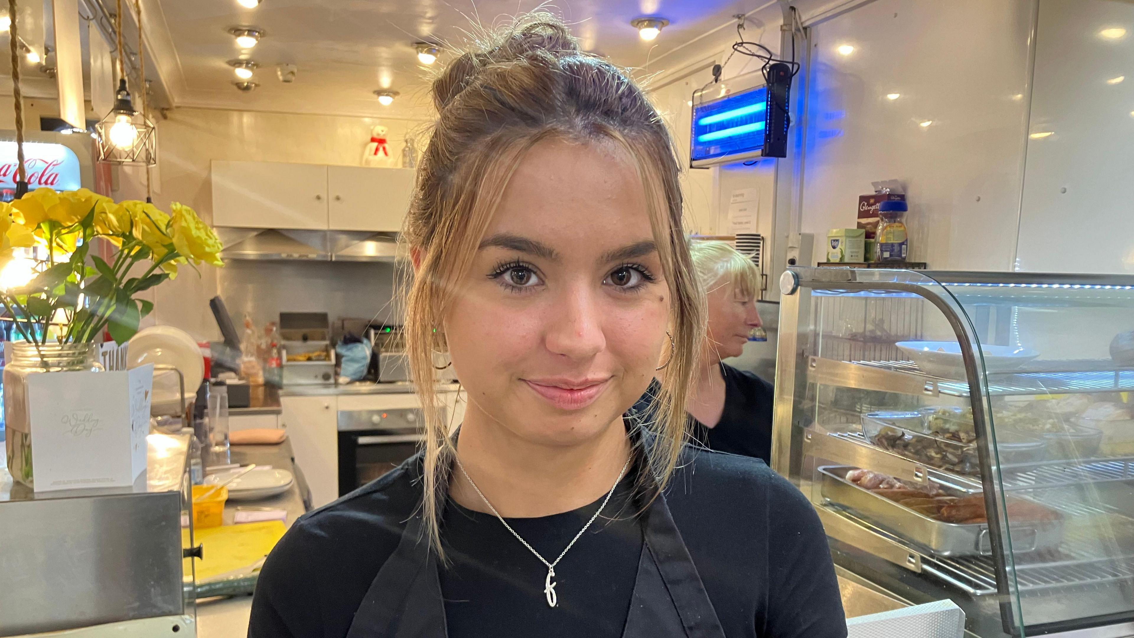 A girl with blonde hair tied back wearing a black top and apron looking at the camera