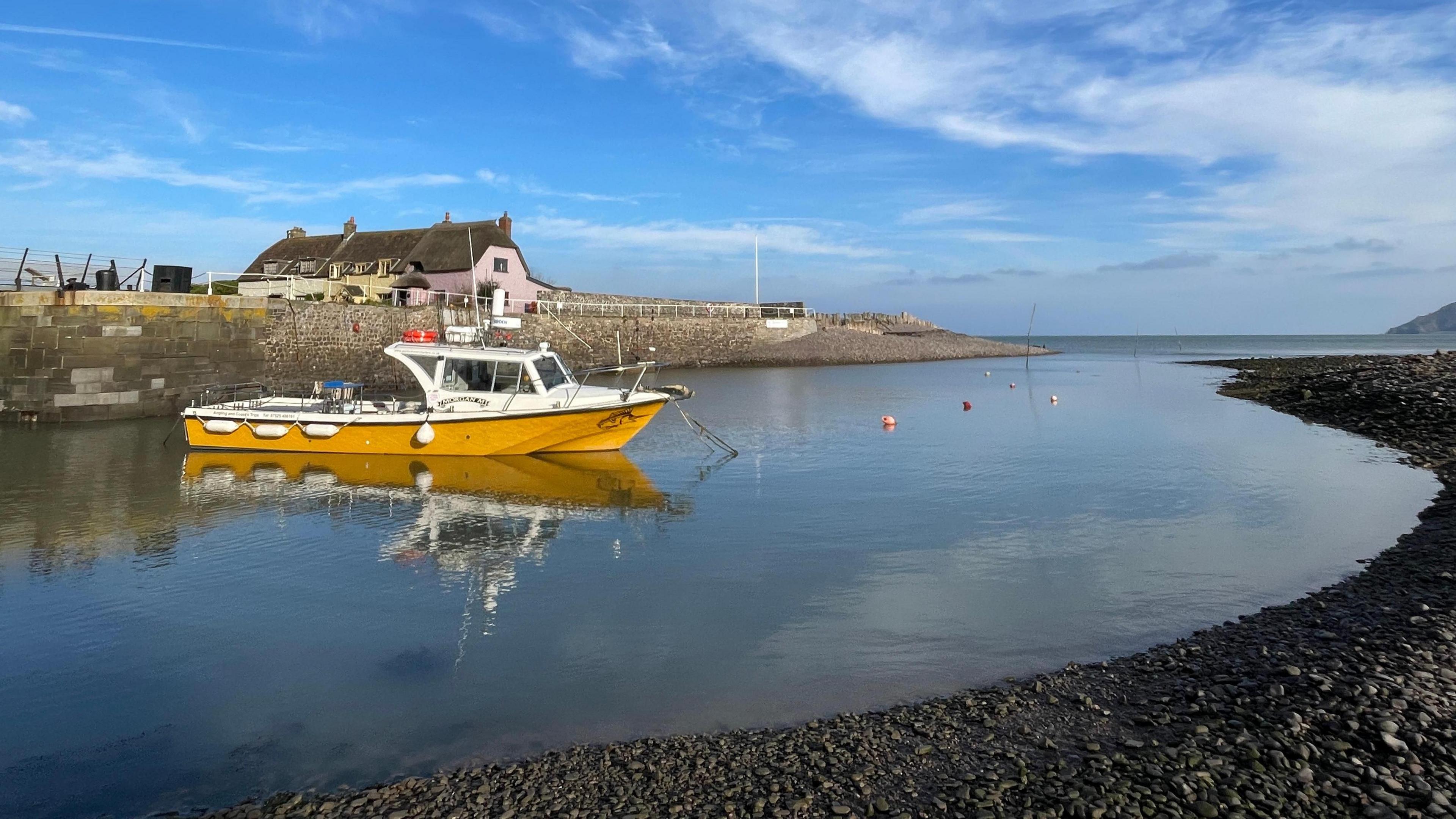 The harbour at Porlock is only half filled with water. A yellow boat is moored and some houses can be seen behind the harbour wall. The sky is blue and mirrored in the water.