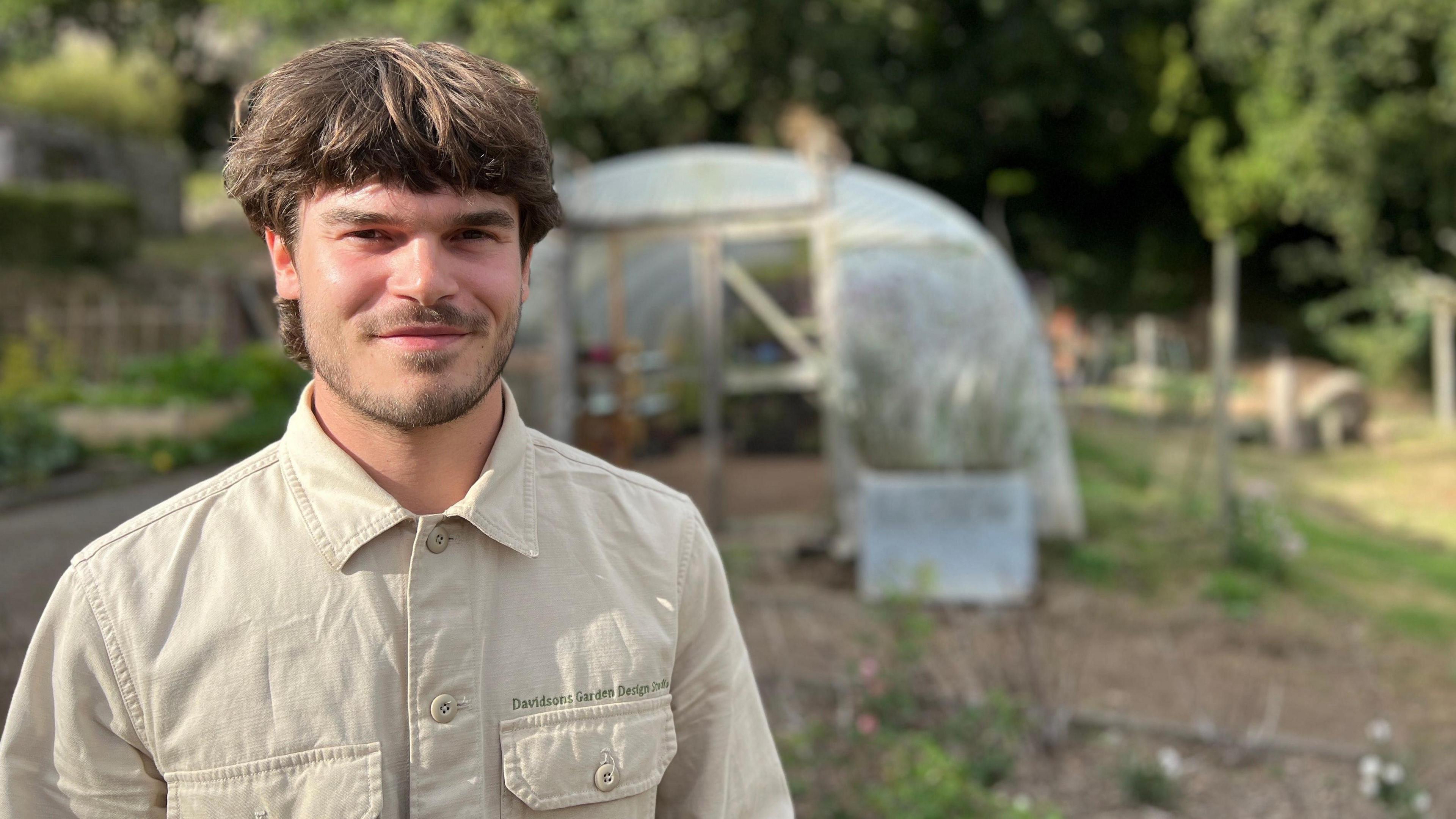 Wills Baker, a man, with dark brown hair and facial hair. He is wearing a beige shirt, buttoned to the top. He is standing in a garden, in front of a greenhouse which is surrounded by greenery.