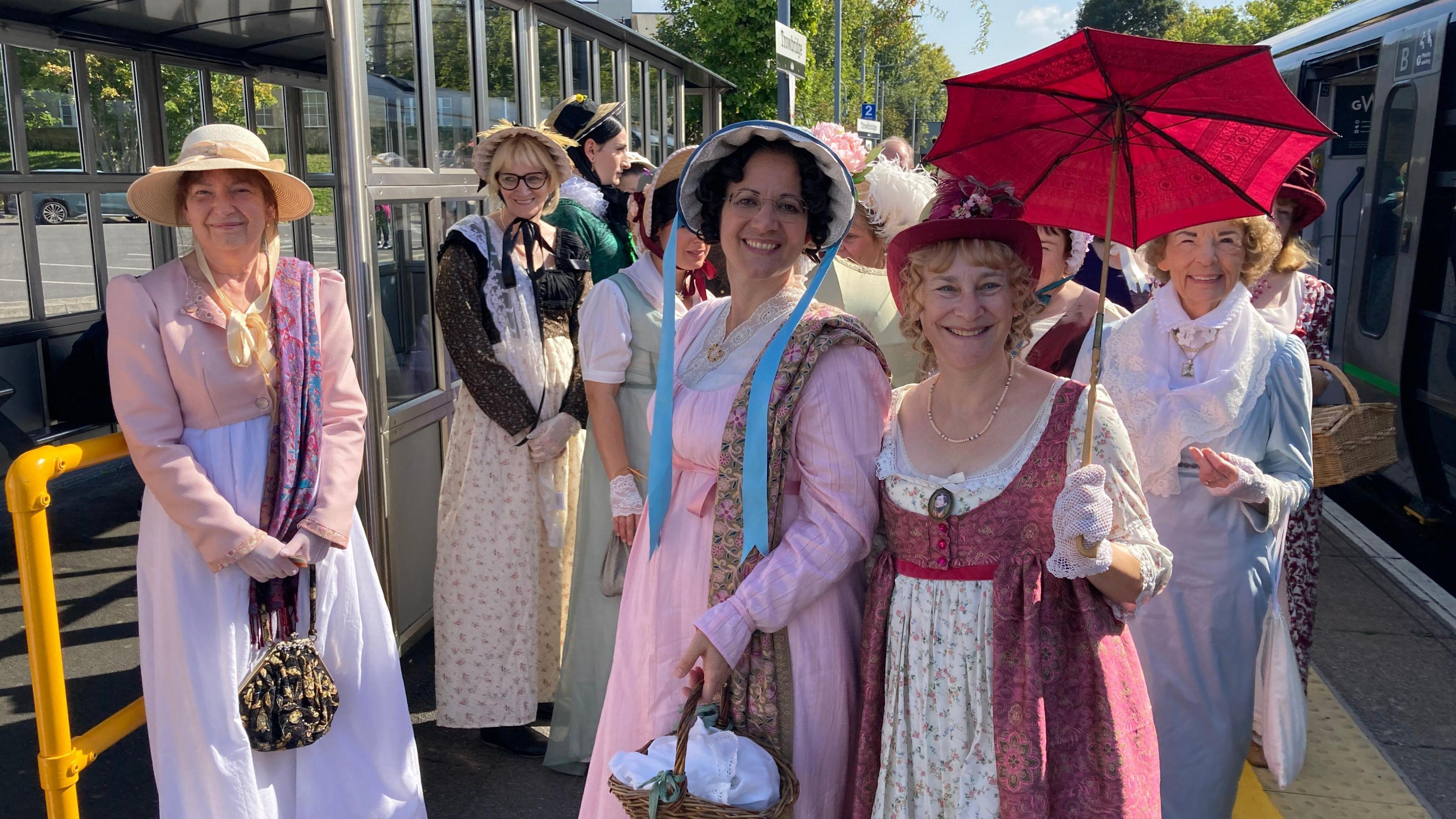 Around 12 women standing on the train platform at Bath Spa station. They are wearing long modest dresses and hats with under the chin ribbons. One is holding a wooden parasol with red fabric, and another is holding a wicker basket. 