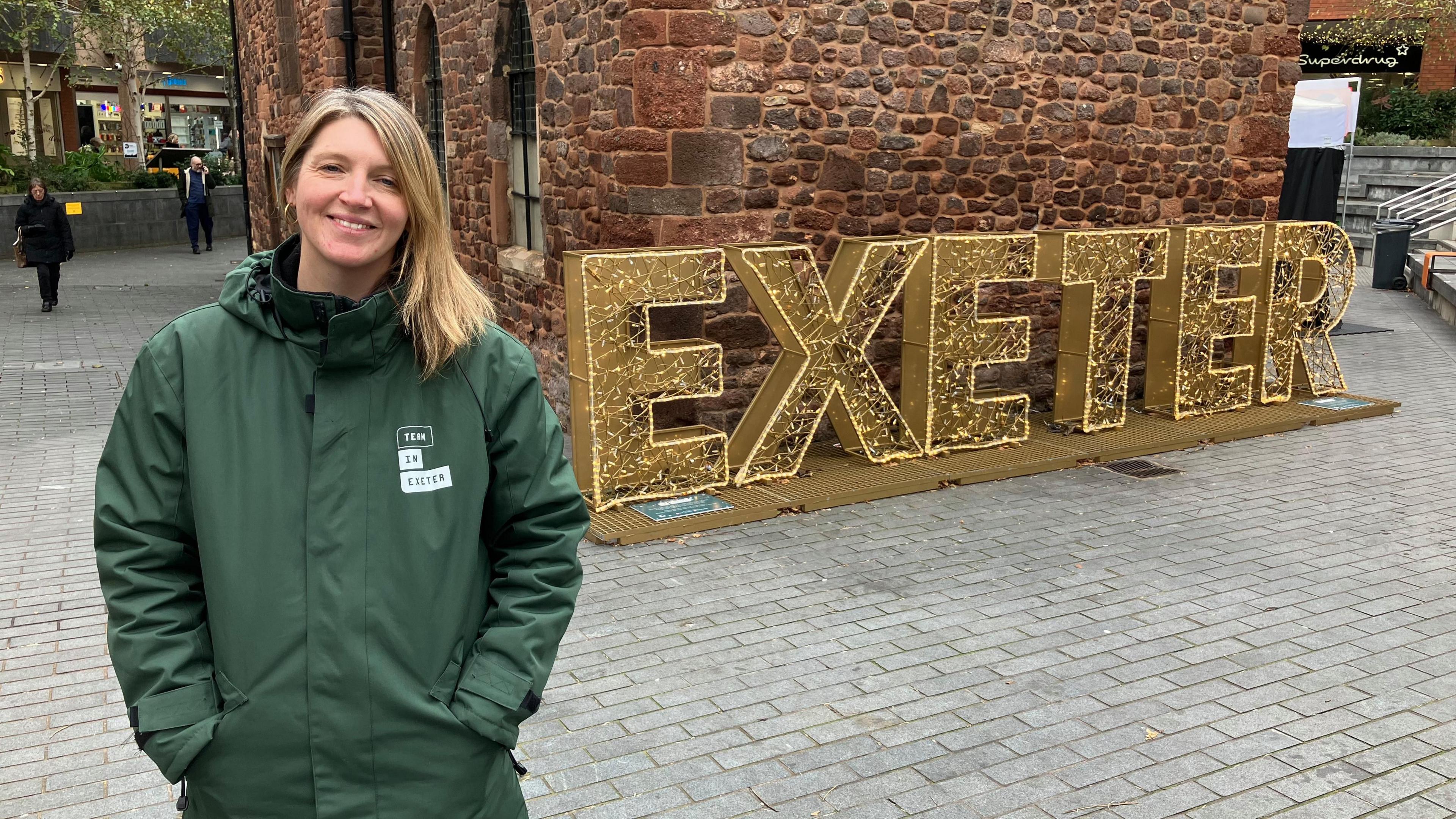 A woman with blond hair wearing a green coat outside a church with an illuminated Exeter sign 