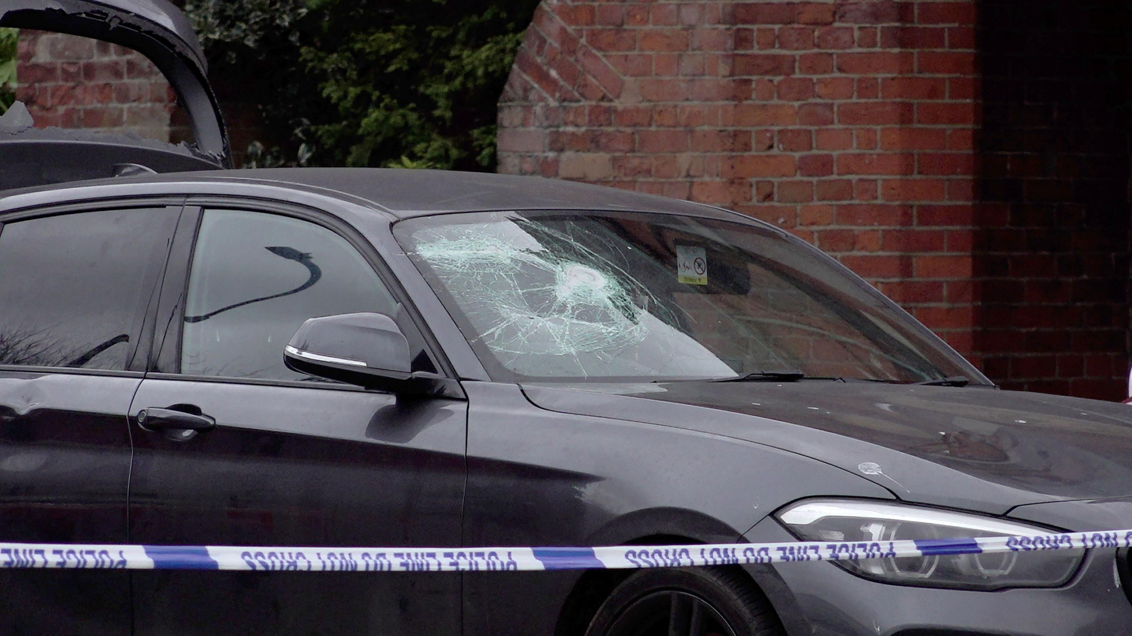 Behind police tape, a grey BMW car windscreen shows cracks and the glass of the open boot is smashed