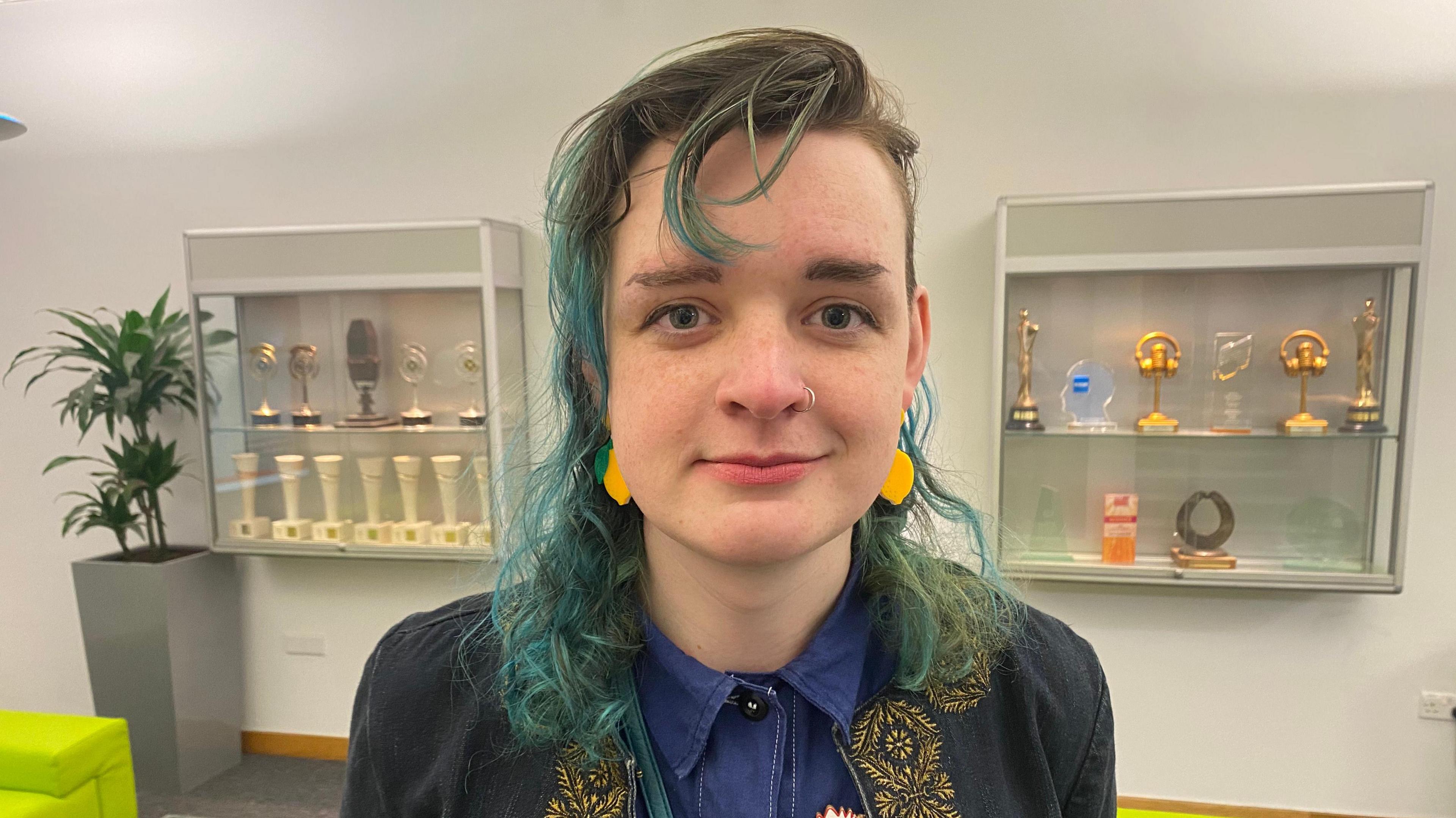 A woman, with dyed blue hair, smiling at the camera. She is pictured from the shoulders up and is wearing a denim shirt and a navy jacket. There is a white wall behind her with two trophy cases. 