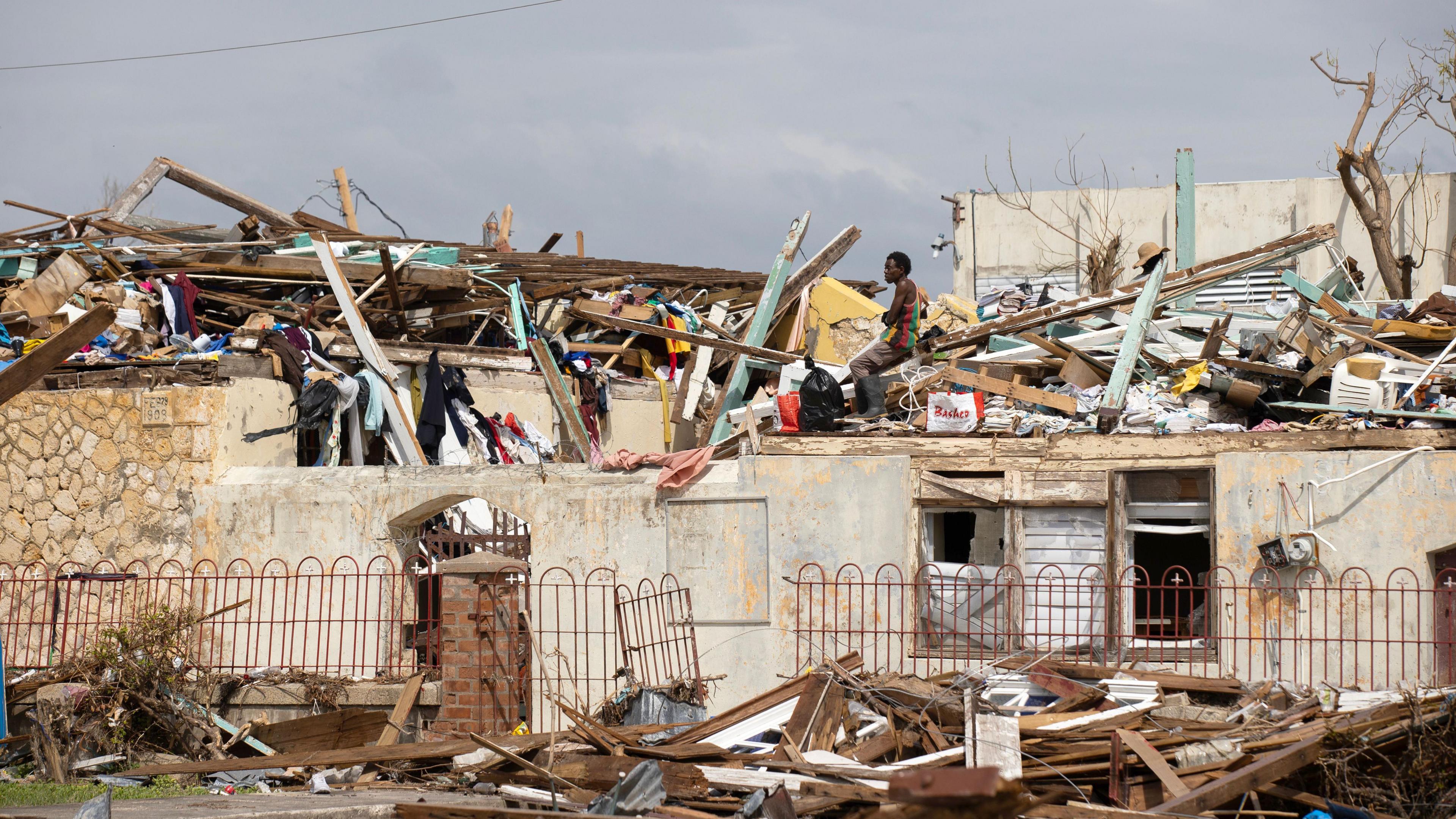 A man searches through the destruction left by Hurricane Melissa in Black River, Jamaica on 11 November. The man is wearing a black, red, green and yellow striped vest and sits on a roof that has been damaged, with piles of planks of wood and other debris sticking out across the rooftops.