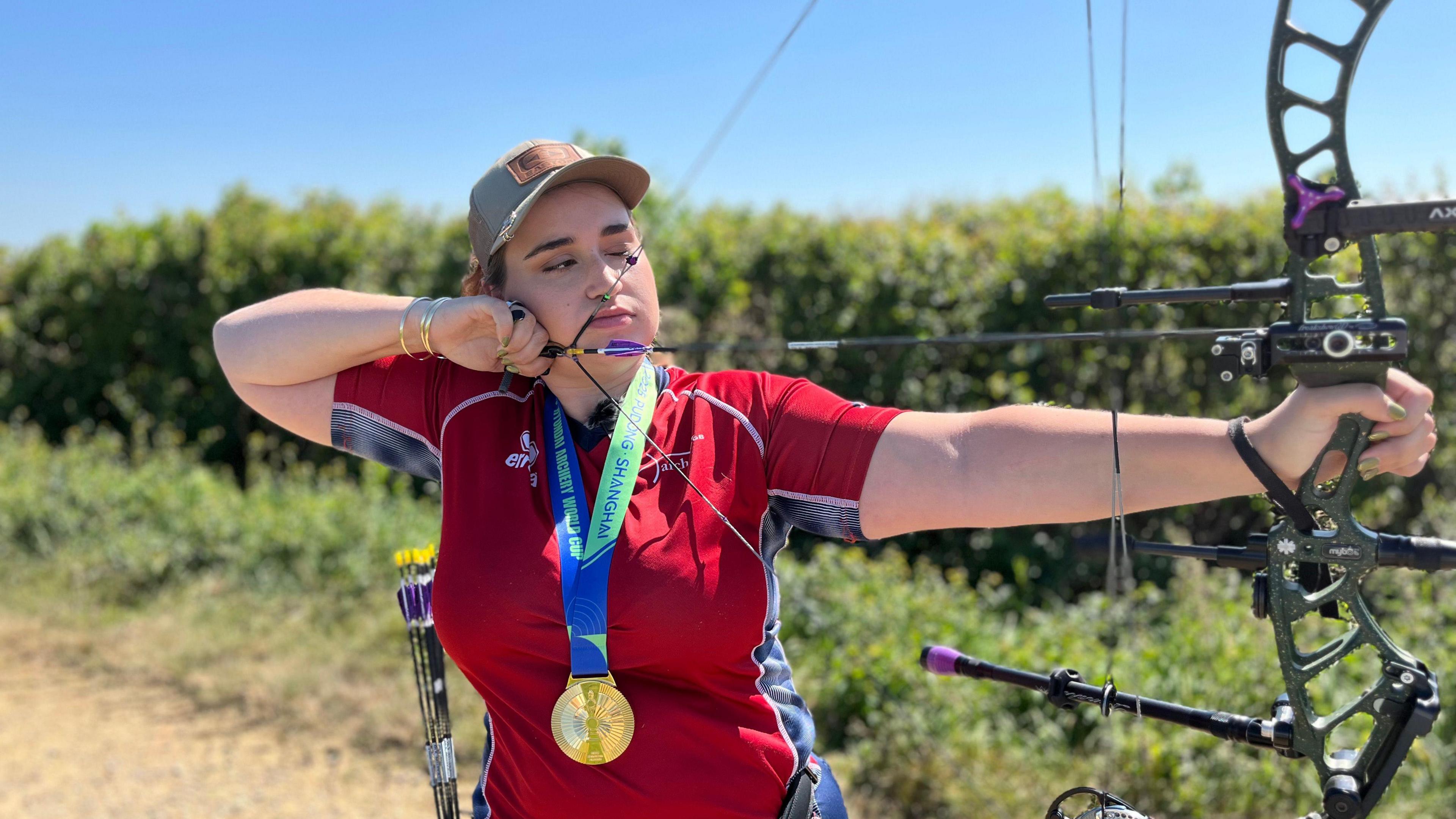 A woman is donning a red sporty shirt and wearing a gold medal around her neck, while pulling a bow that looks very sophisticated. She looks very concentrated as if she's about to shoot an arrow. She has a small number of arrows behind her waste ready to use next.