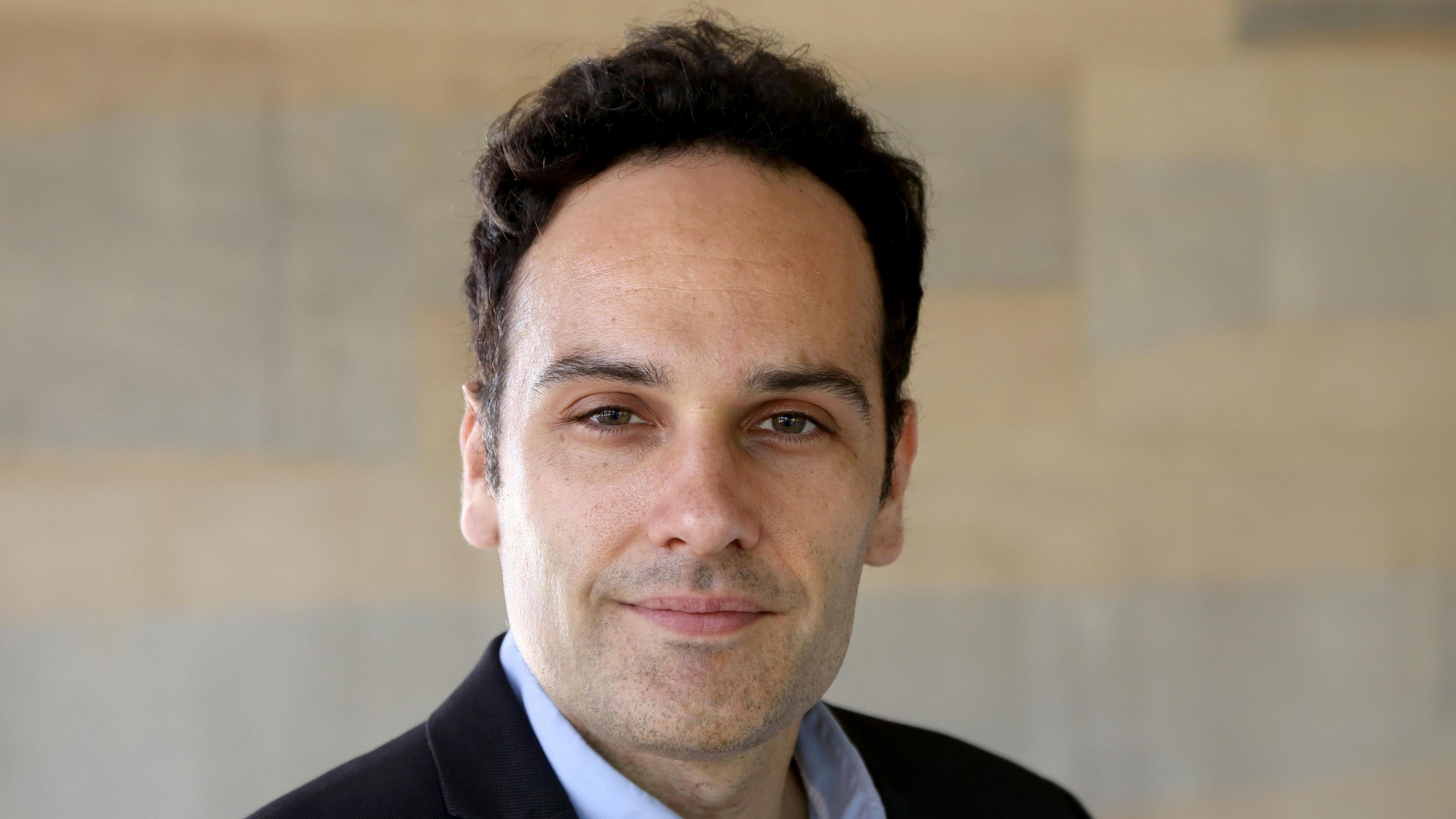 Dr Javier Andreu-Perez is smiling. He has black hair and stubble. He is wearing a black suit jacket and a pale blue shirt. There is a wall behind him, although it has been blurred.