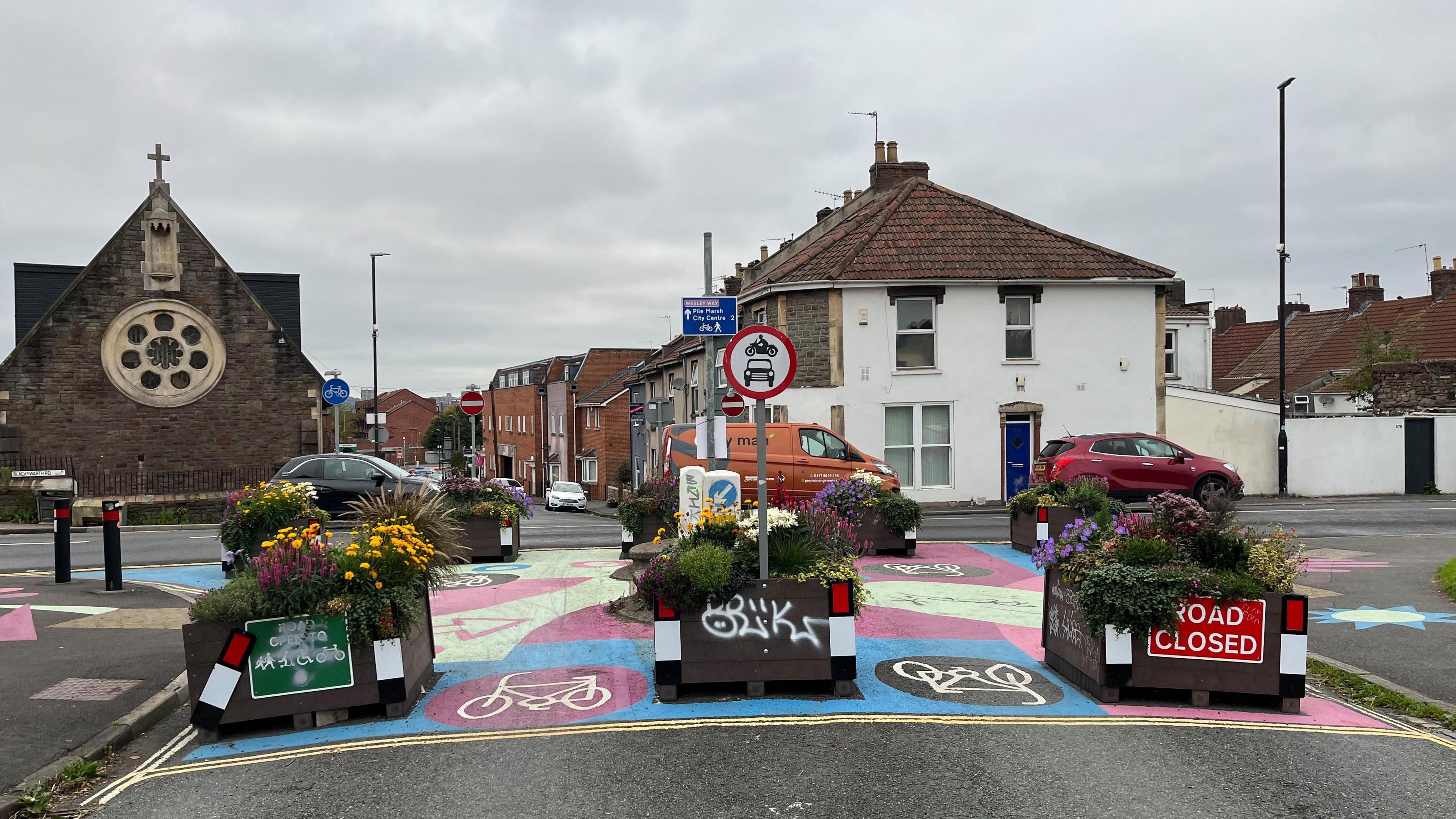 A row of planters blocking the end of a road, with space for bicycles to pass between them. One of the planters has a "road closed" sign on it. On the other side is a queue of traffic heading along a main road.